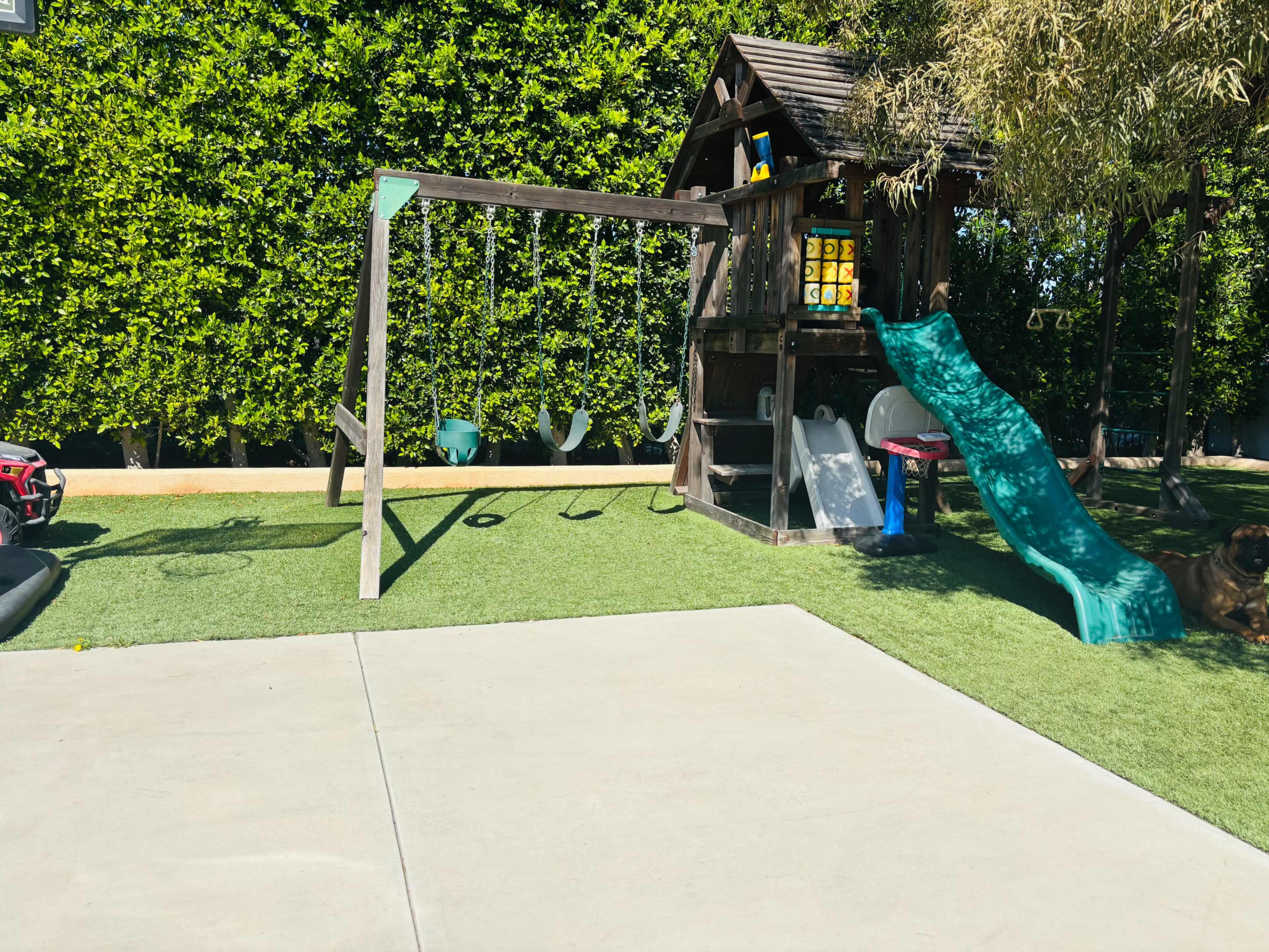 The image shows a playground with a wooden play structure, swings, a green slide, and artificial turf surrounding a concrete area.