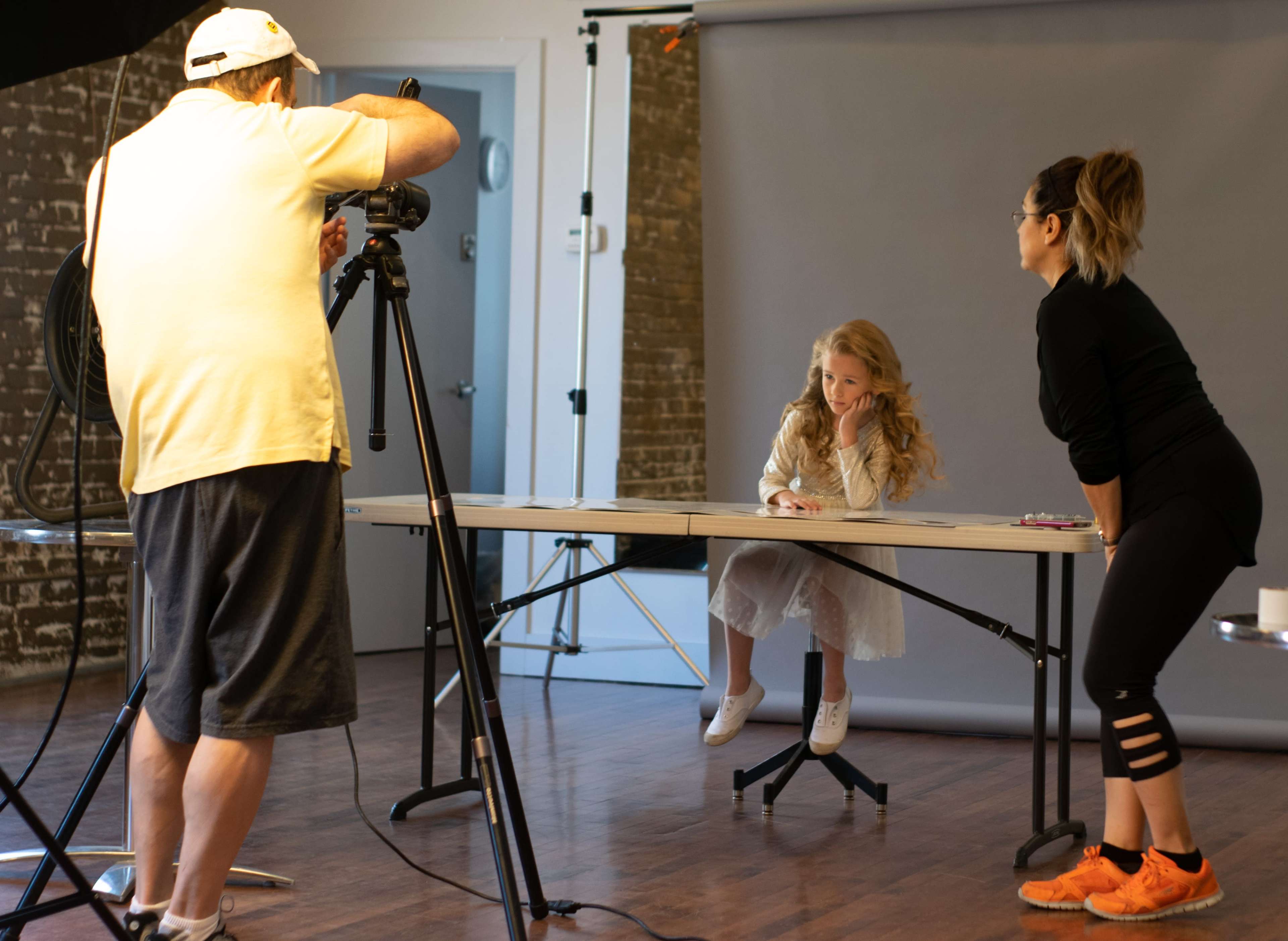 A photographer captures a young girl seated at a table while a woman assists off to the side in a well-lit studio.