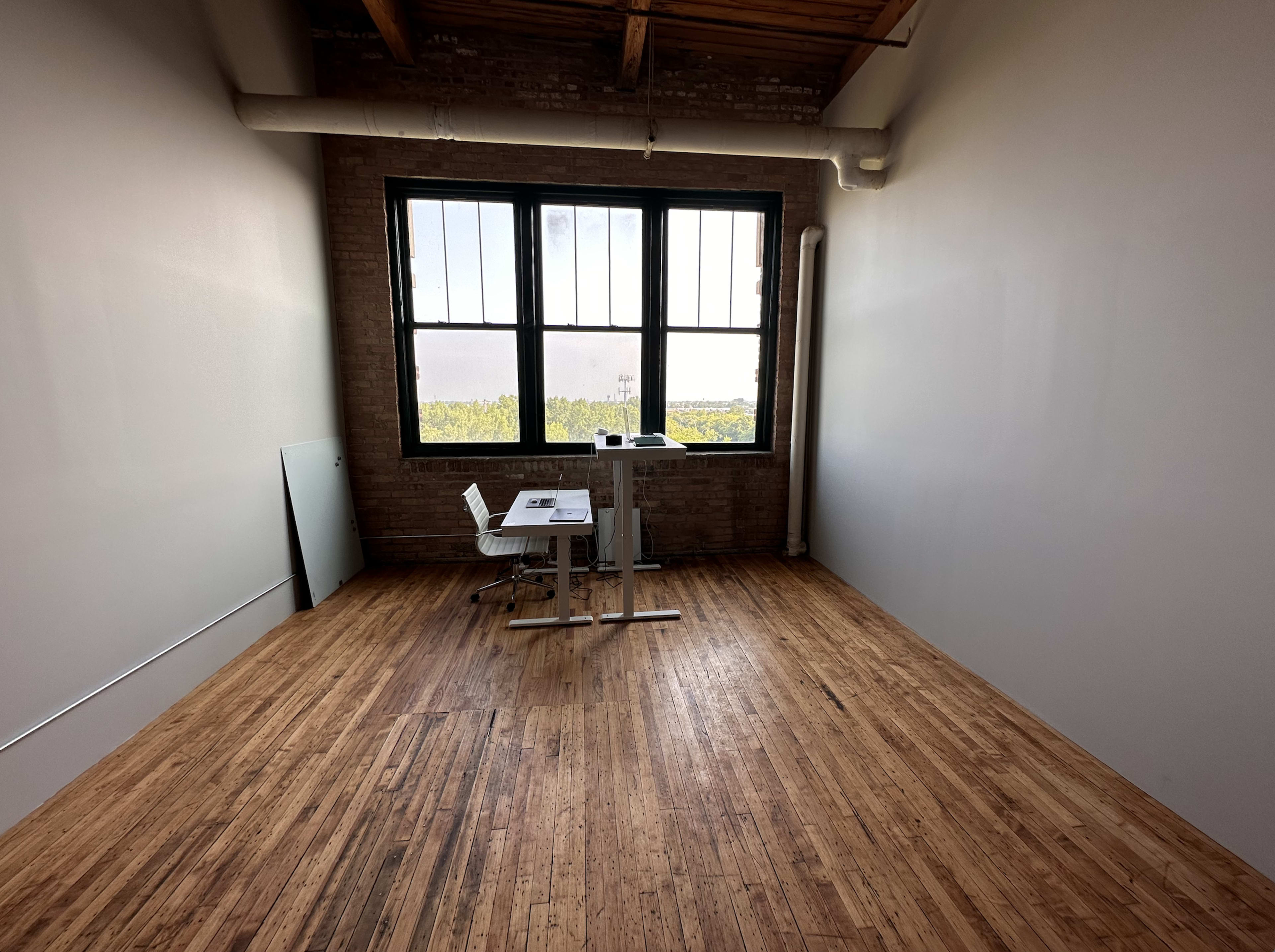 A sparse office space with wooden floors, a white table and chair, and large windows overlooking a green landscape.