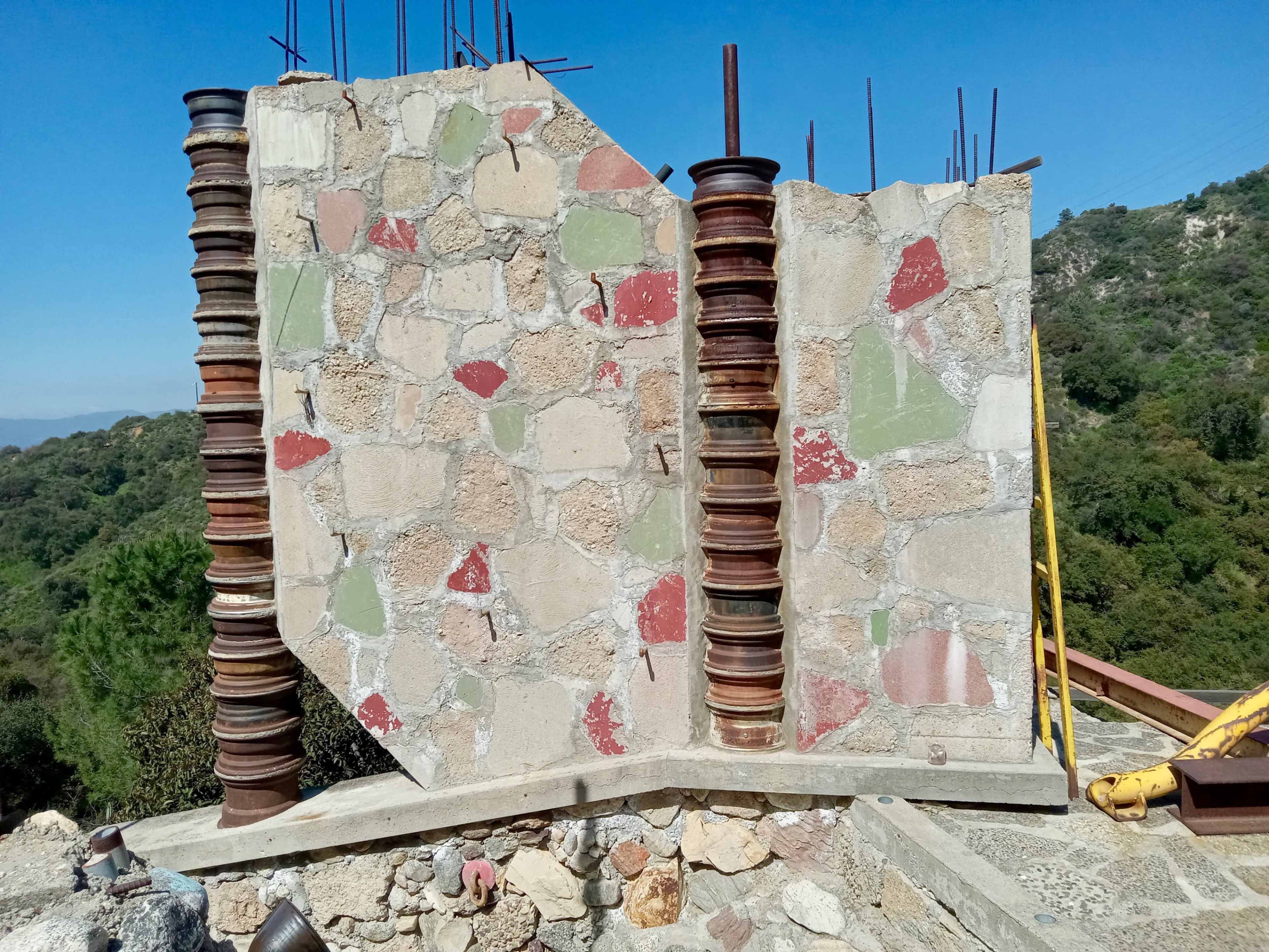 The image shows a partially constructed stone wall with exposed metal reinforcements and colorful patches, set against a landscape of trees and hills under a clear blue sky.