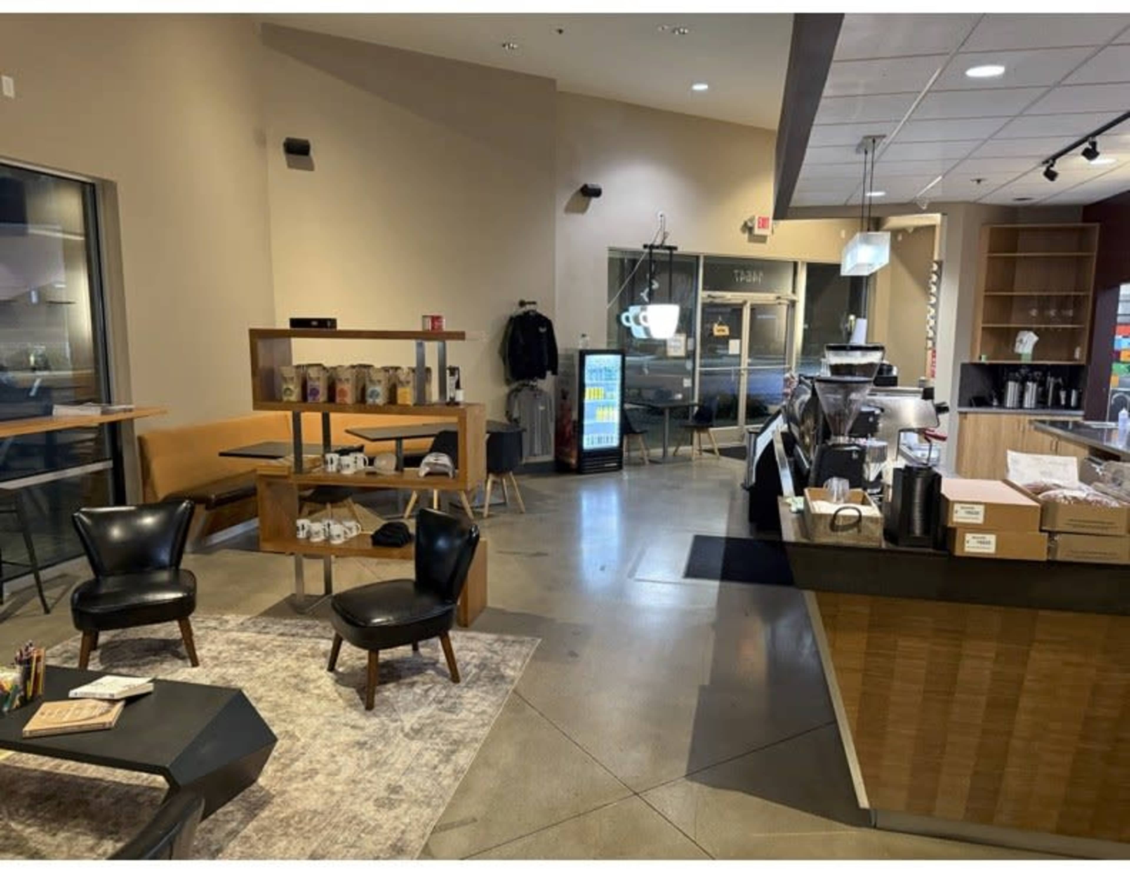 The image shows a modern café interior with black chairs, a wooden display shelf, a coffee counter, and a visible menu board.