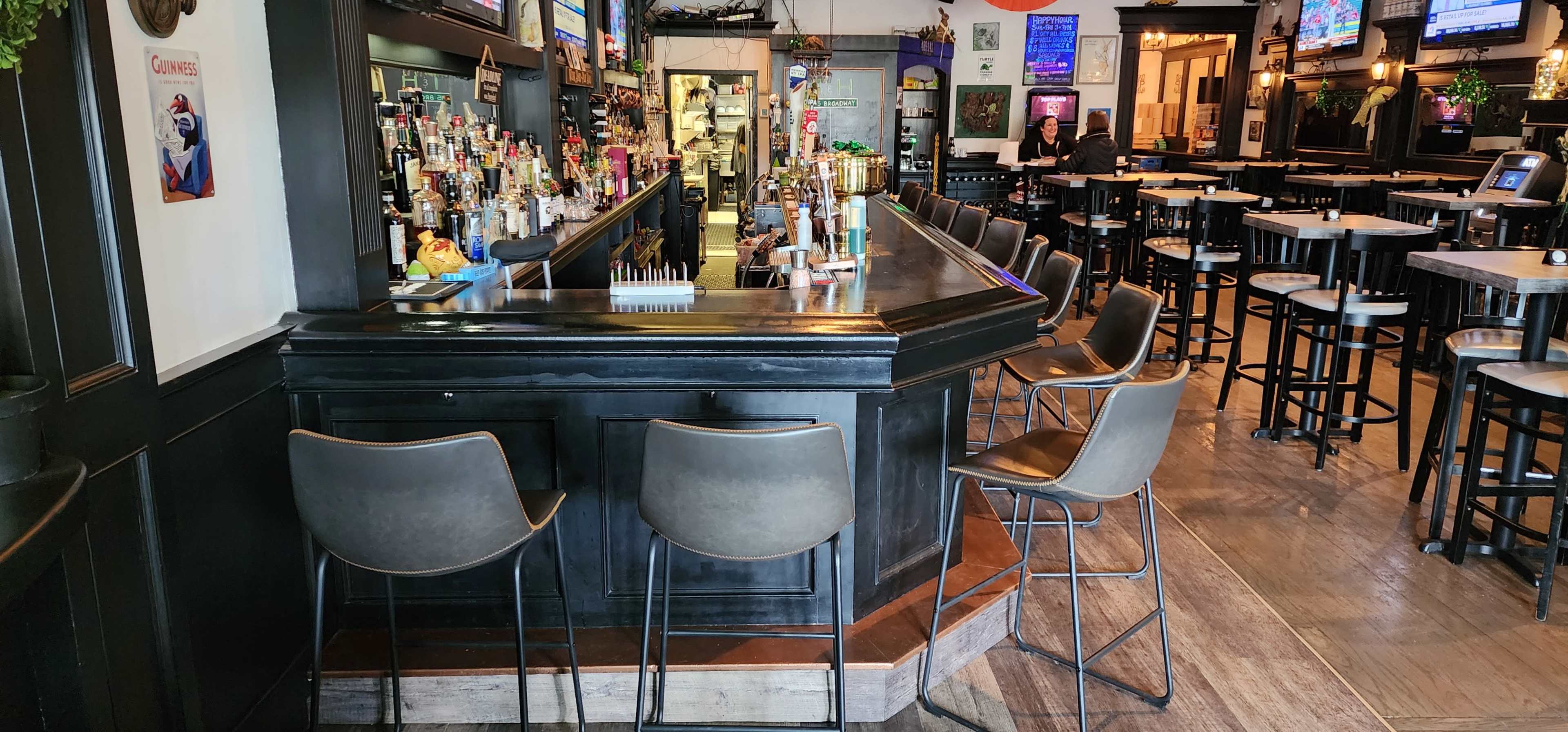 A well-lit bar area with a long counter and several high stools, surrounded by tables and chairs in a casual dining space.