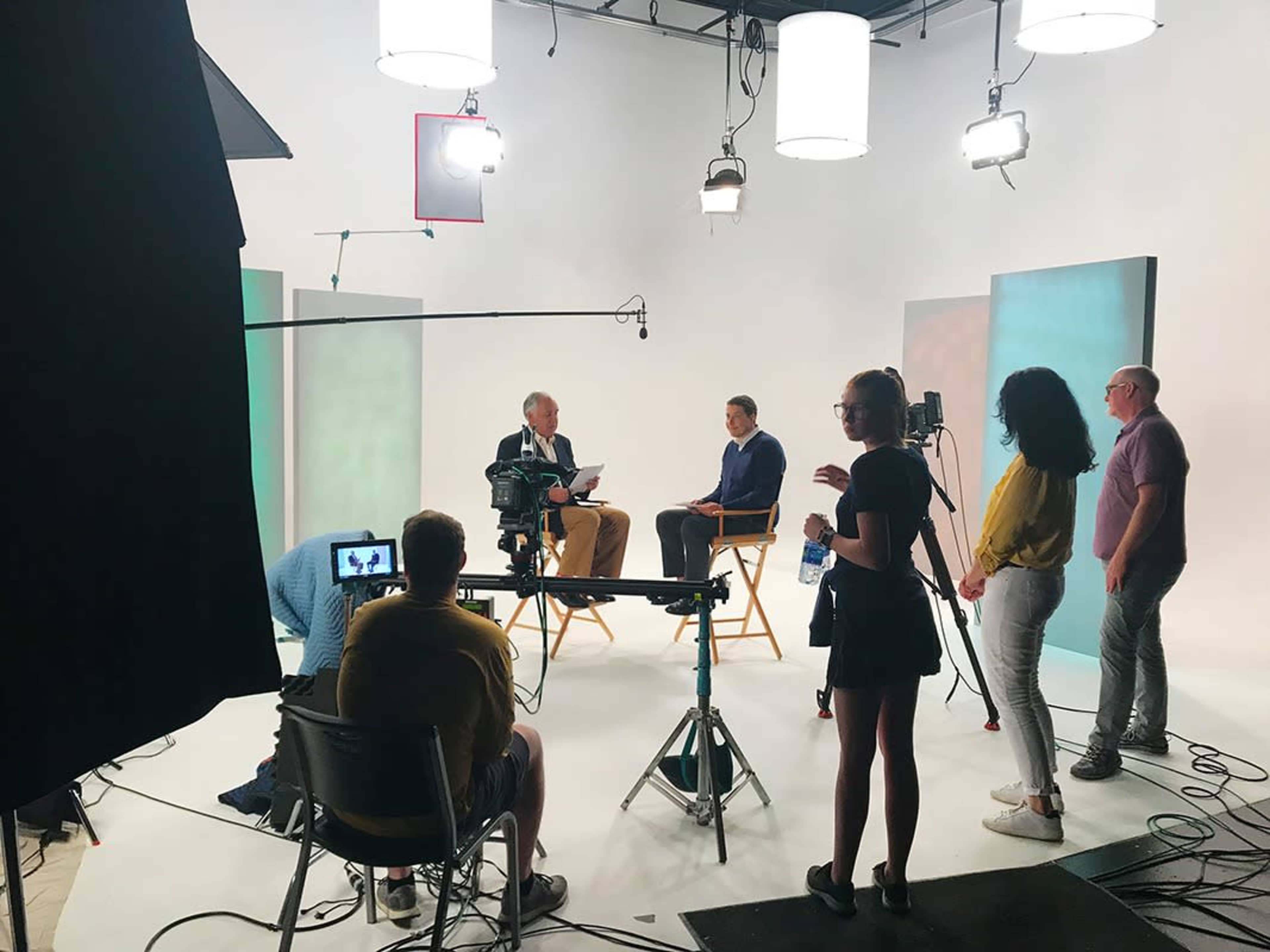 A television studio set is arranged for an interview, featuring multiple cameras, lighting equipment, and crew members preparing around two seated guests.