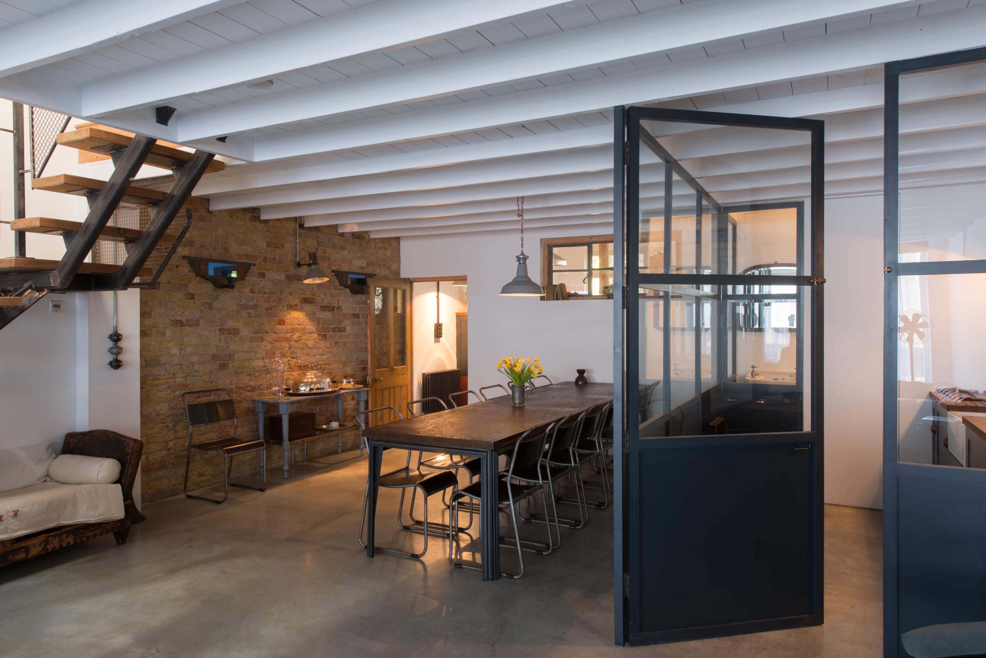 A modern interior space featuring a dining area with a long table, a kitchen area visible through glass partitions, and a staircase with wooden steps against a brick wall.