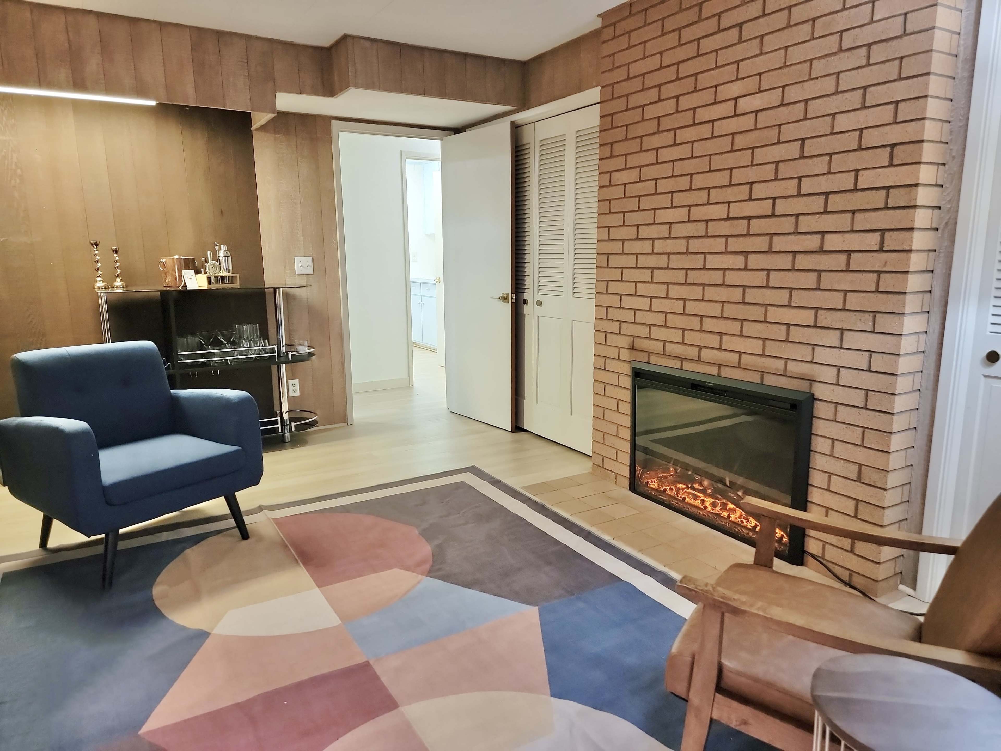 The image shows a cozy room featuring a blue armchair, a brown wooden rocking chair, a modern electric fireplace, and a patterned area rug, with a bar cart in the corner and a doorway leading to another room.