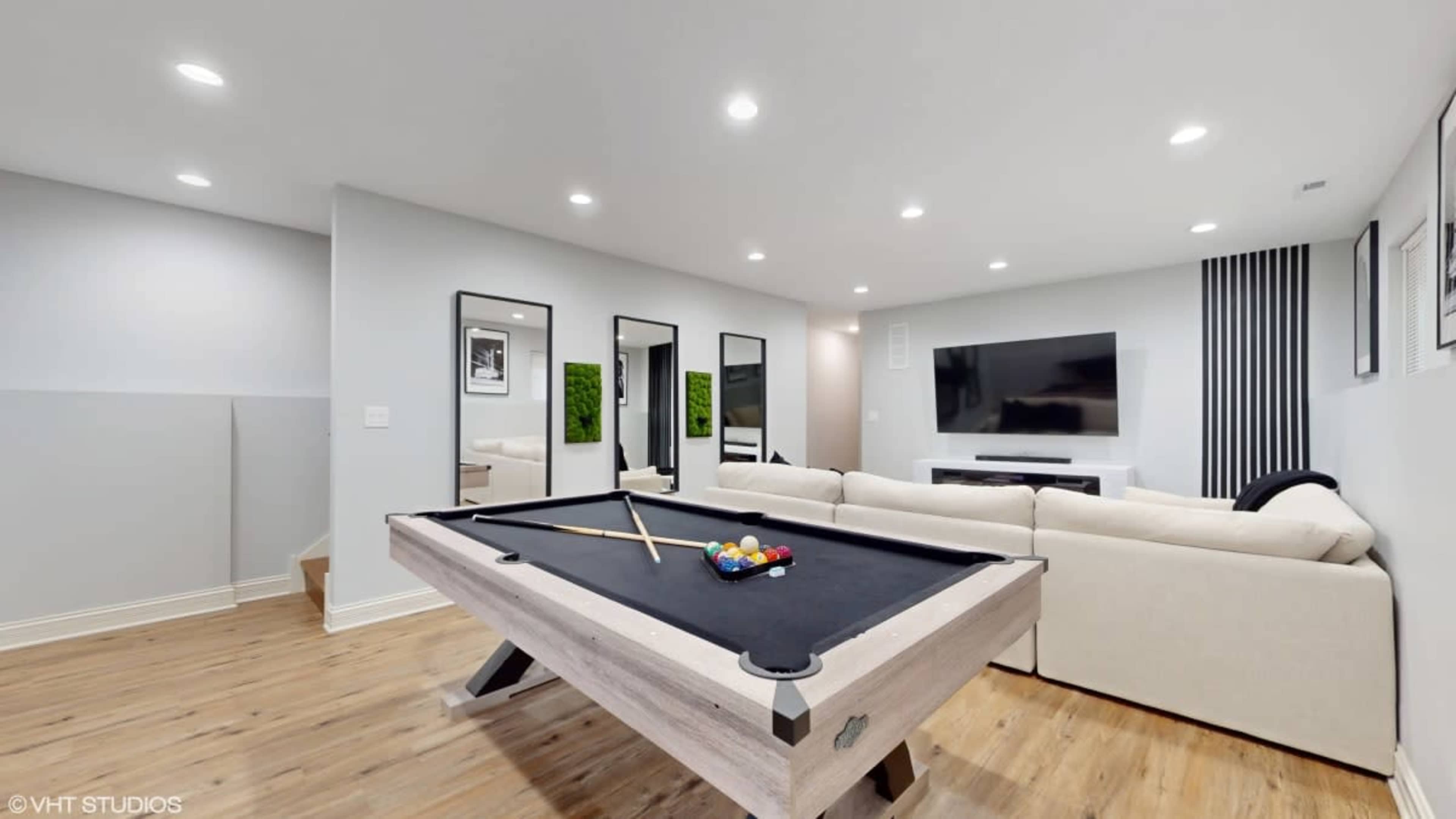 The image shows a modern basement recreation room featuring a pool table, sectional sofa, and a large wall-mounted television.