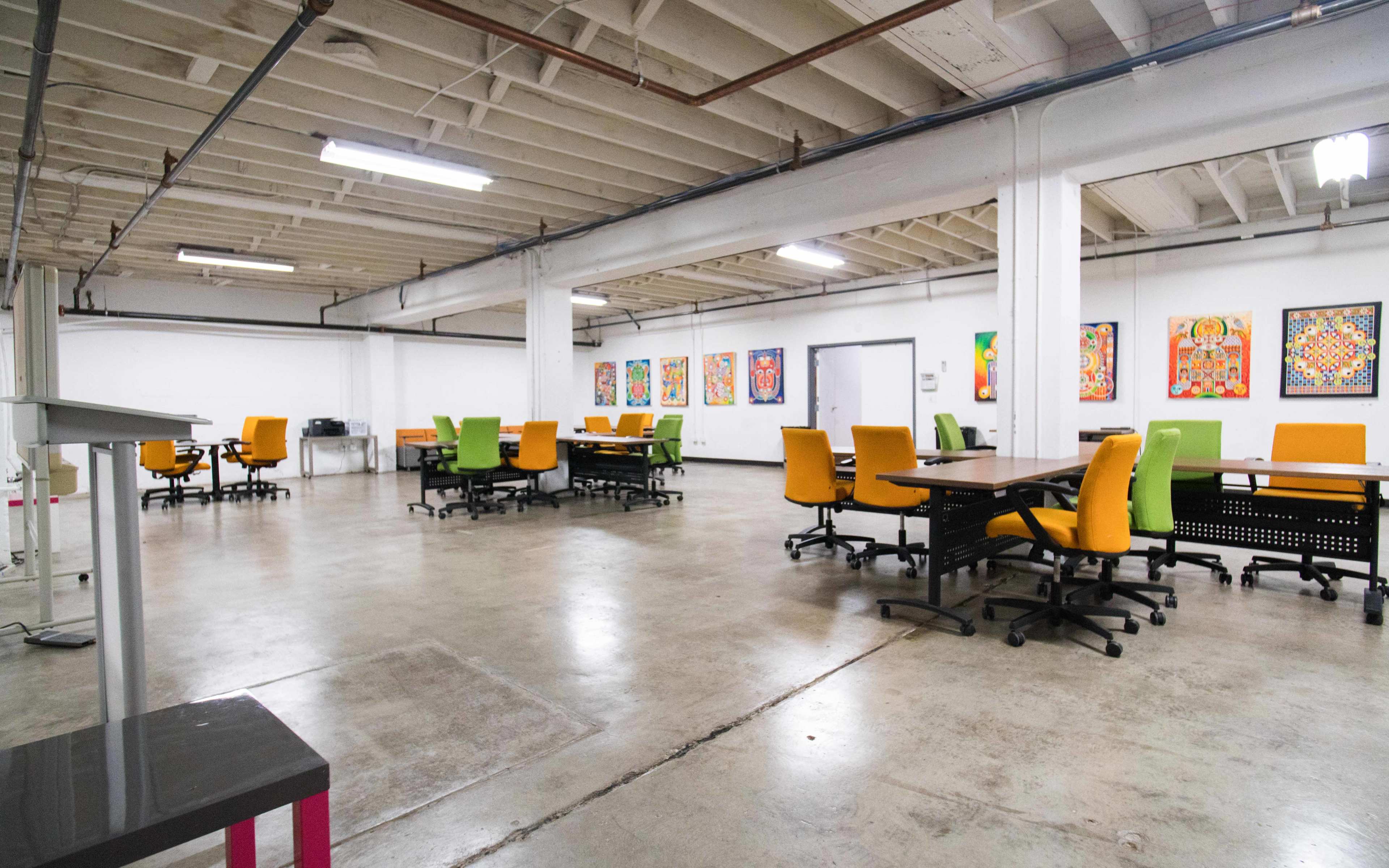 A spacious workplace with multiple tables and chairs, colorful artwork on the walls, and a concrete floor.