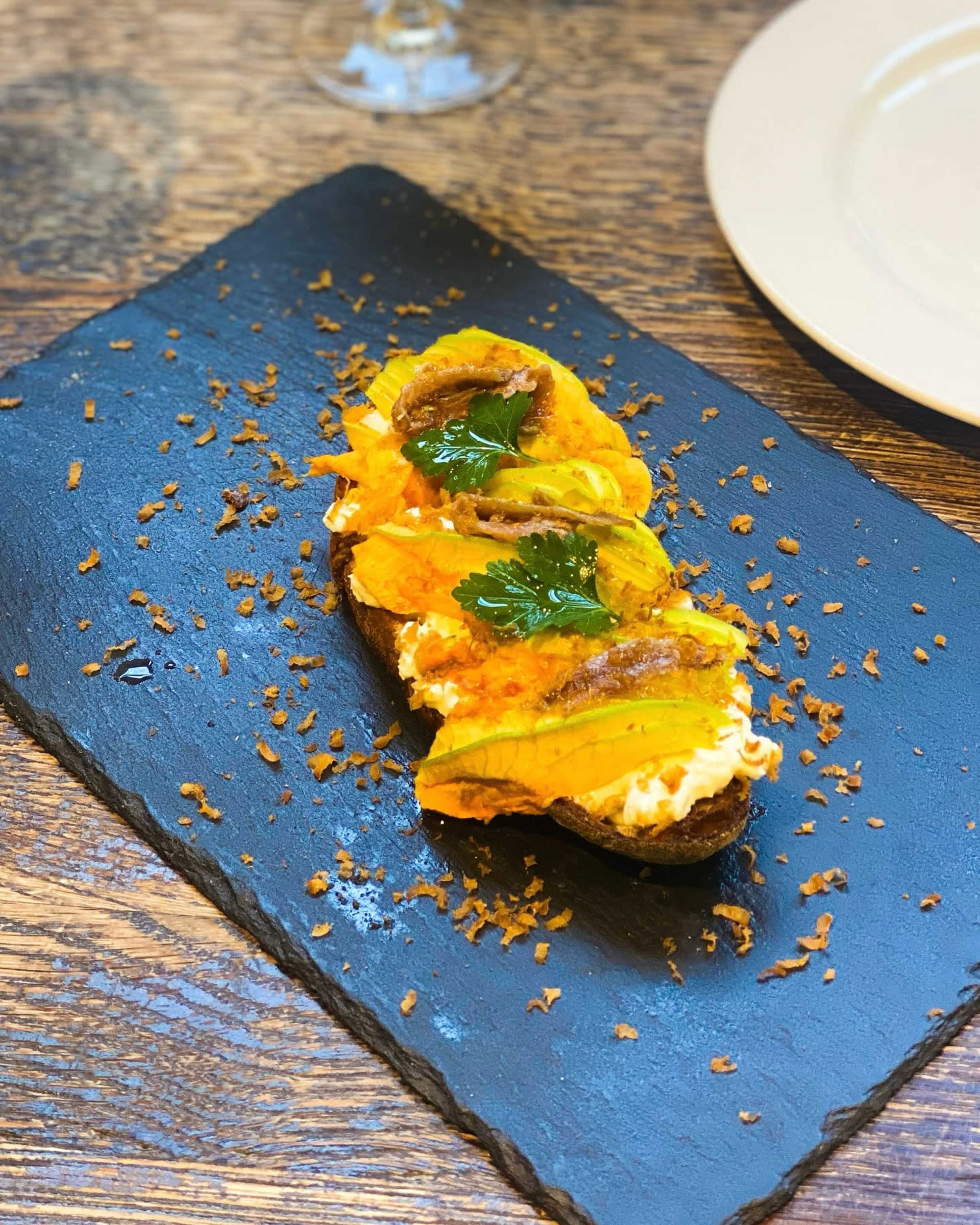 A slice of dark bread topped with layers of colorful vegetables, cream, and garnished with a sprig of green herb, served on a black slate plate.