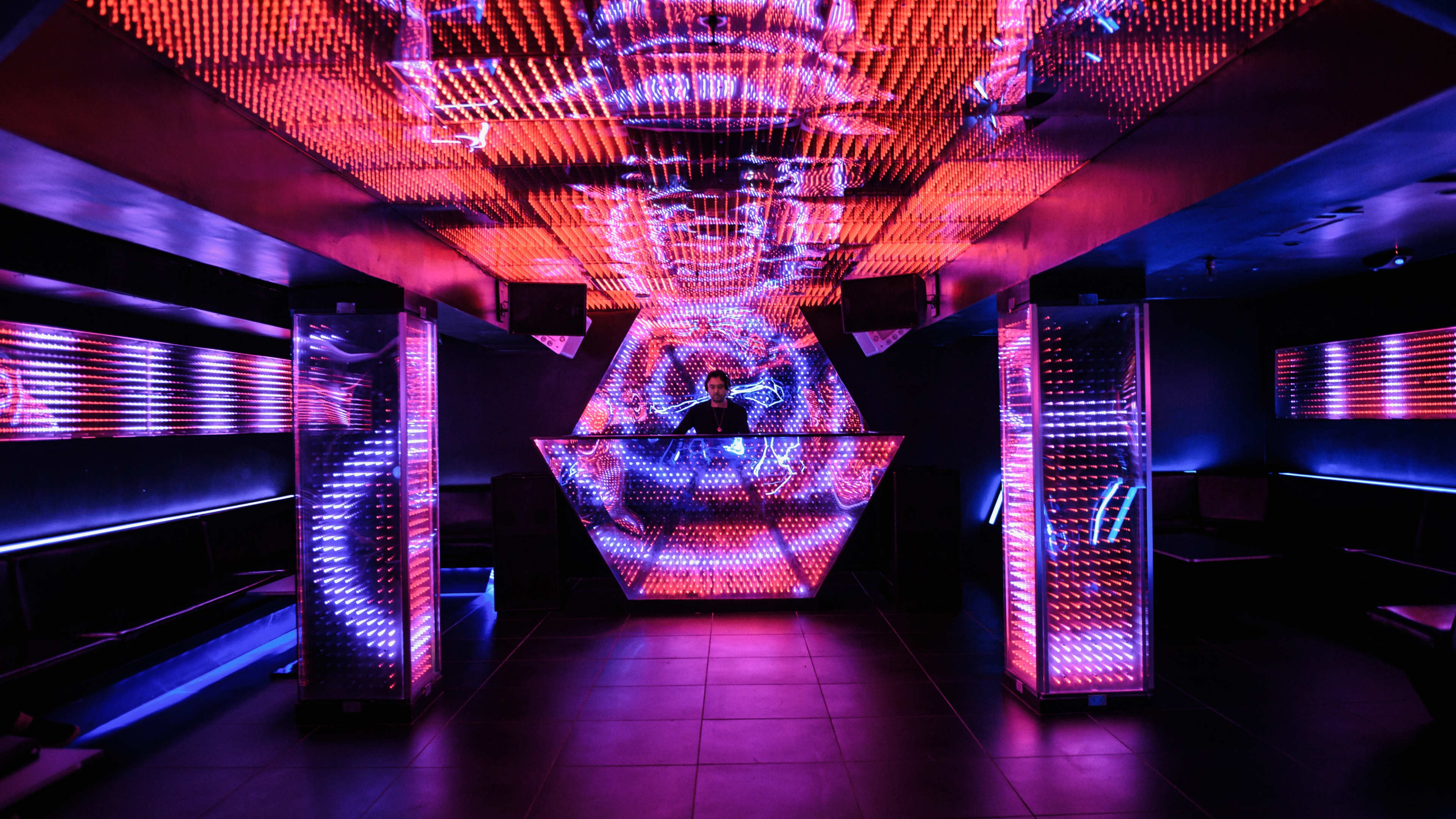 A DJ stands behind a neon-lit hexagonal booth in a vibrant nightclub filled with colorful LED lights.