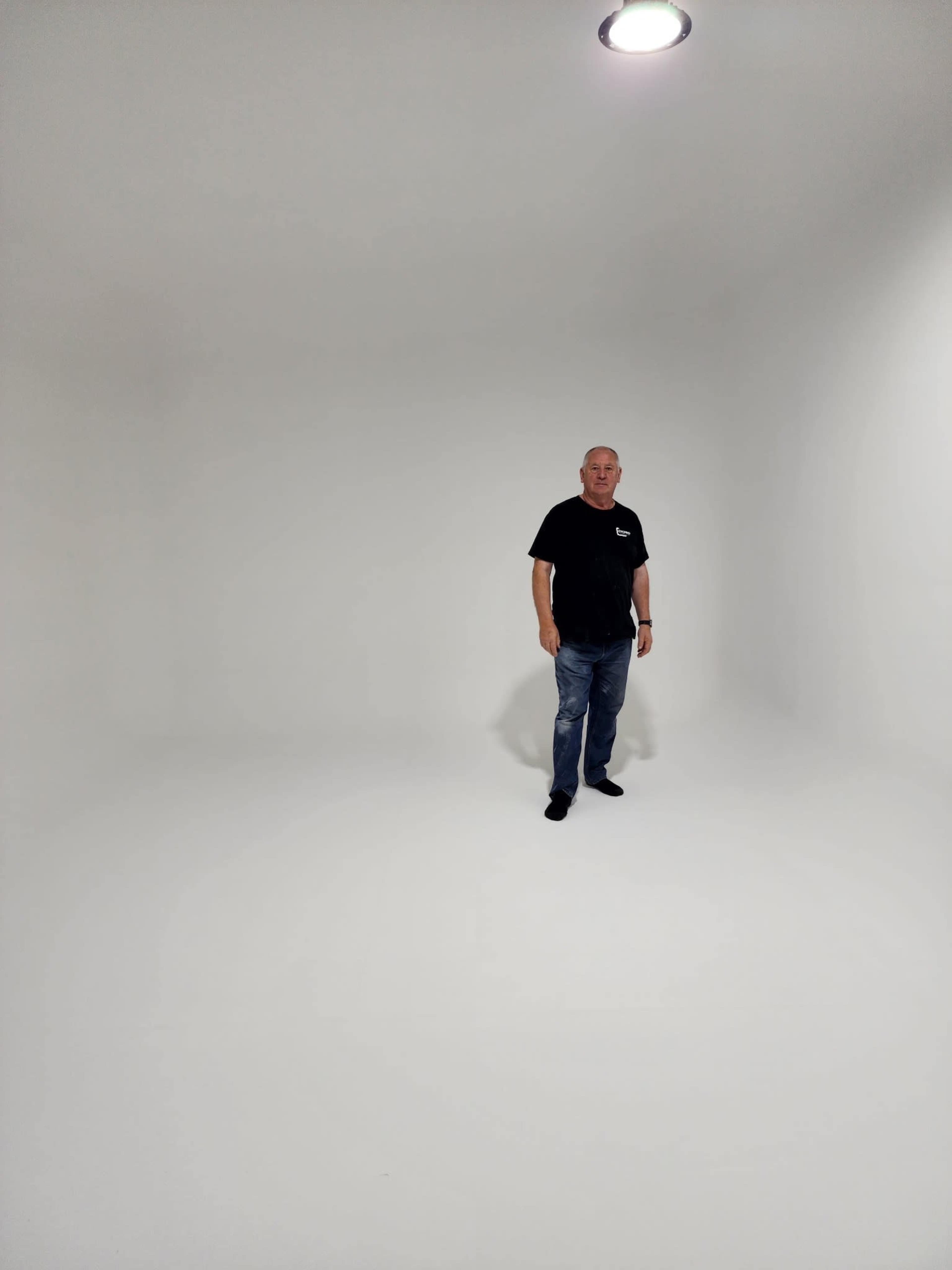 A man stands in a light-colored, empty room with smooth walls and a single overhead light.