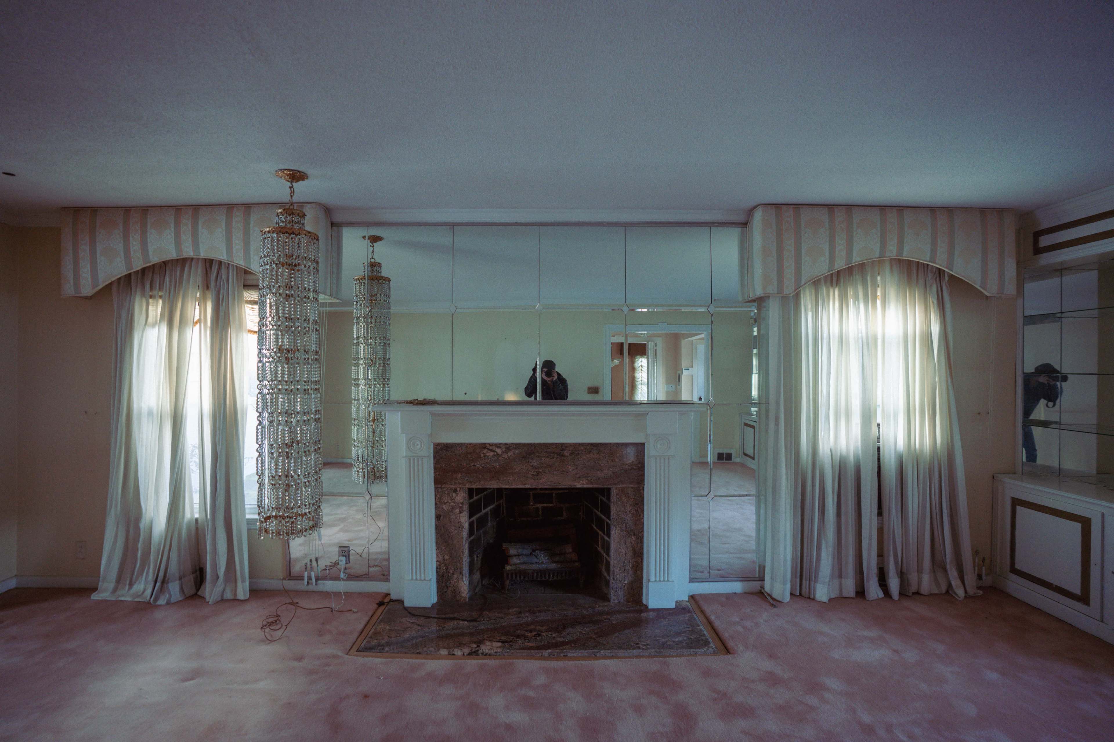 The image shows a spacious living room featuring a mirrored wall, a fireplace with a marble surround, and long, sheer curtains draped beside two hanging chandeliers.