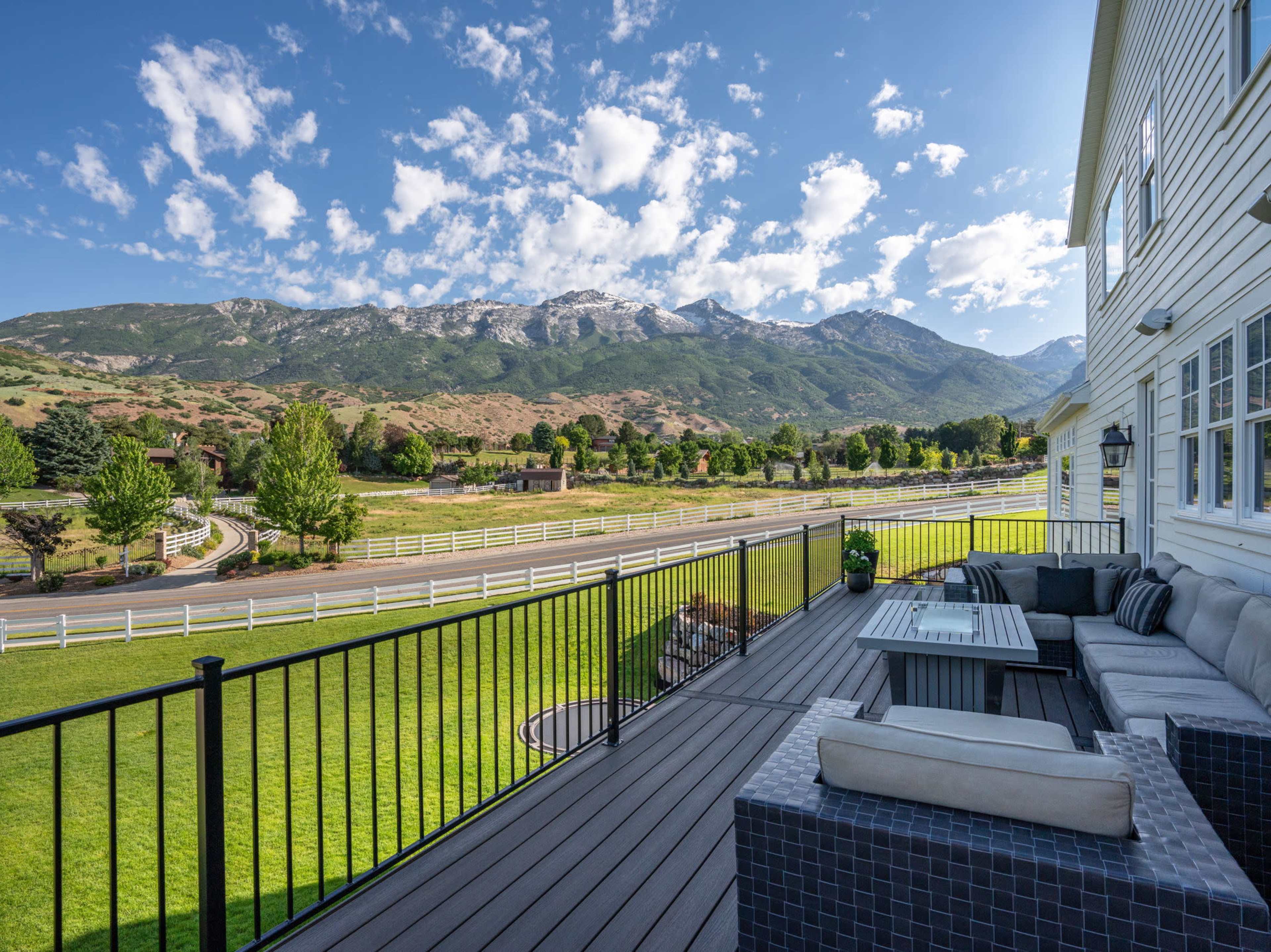 A spacious wooden deck with a seating area overlooks a green lawn and mountainous landscape under a partly cloudy sky.