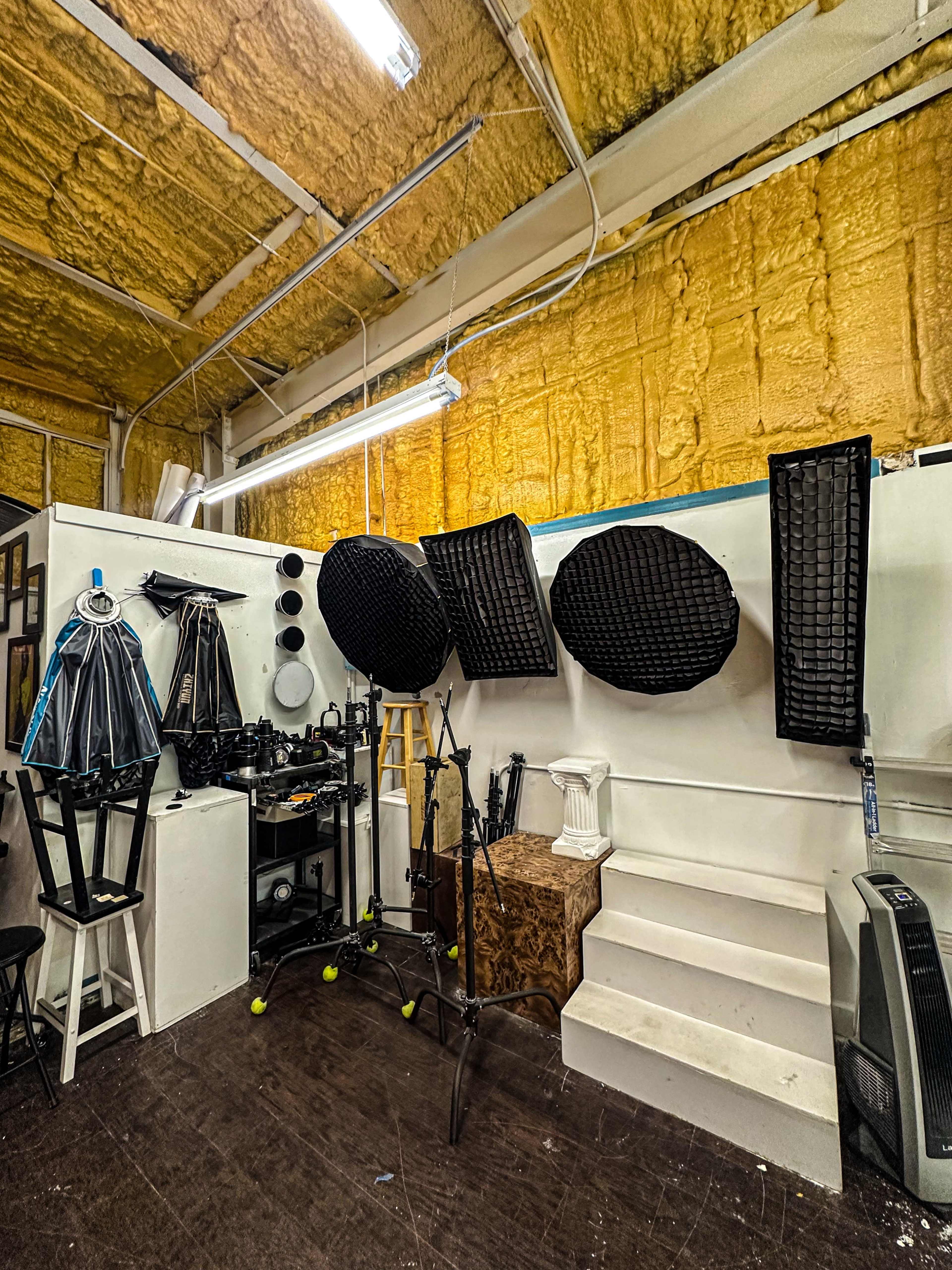 The image shows a photography studio with various lighting equipment, backdrop materials, and a set of stairs against a wall with insulation.