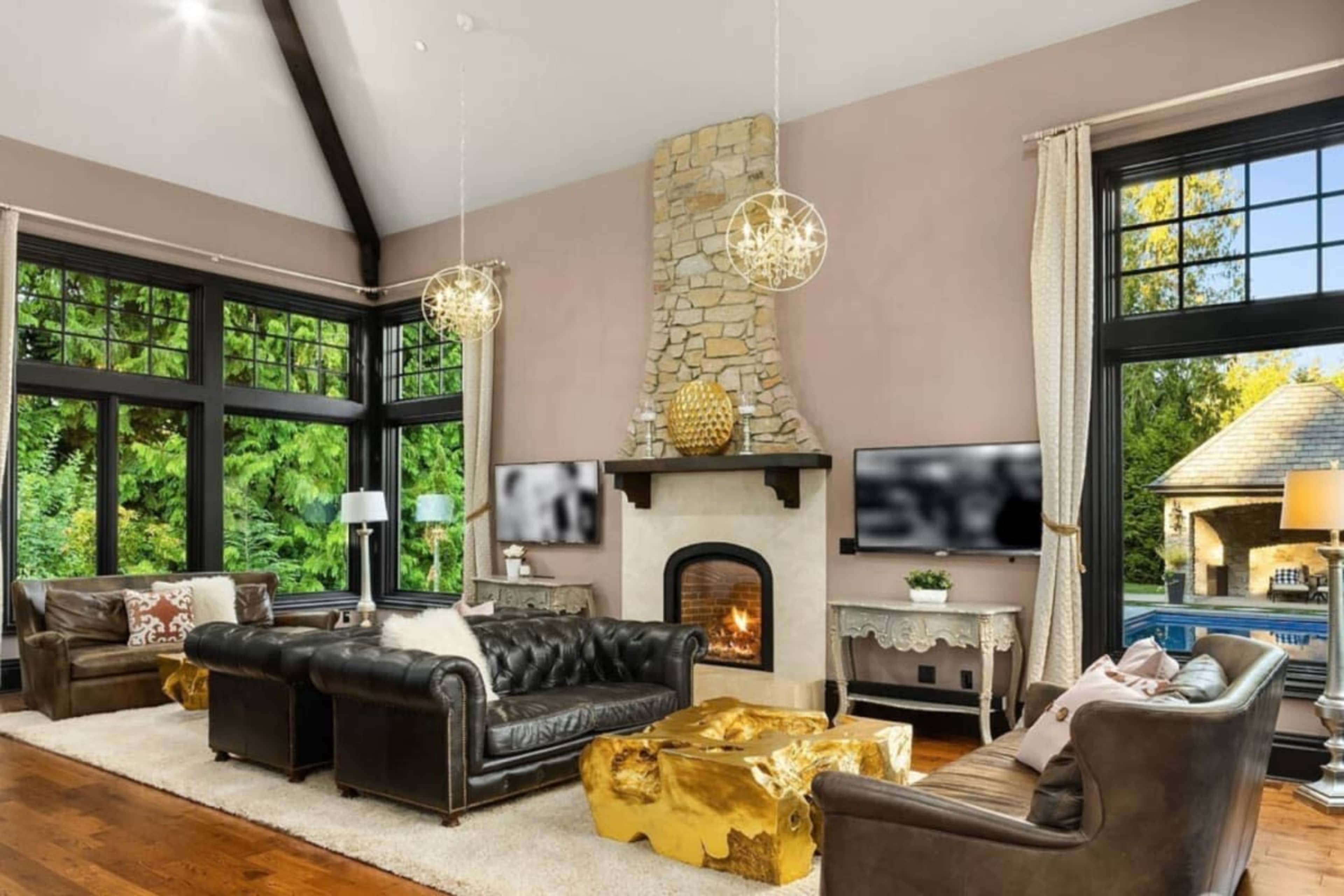 The living room features a stone fireplace, large windows with views of greenery, and a mix of leather furniture and wooden accents.