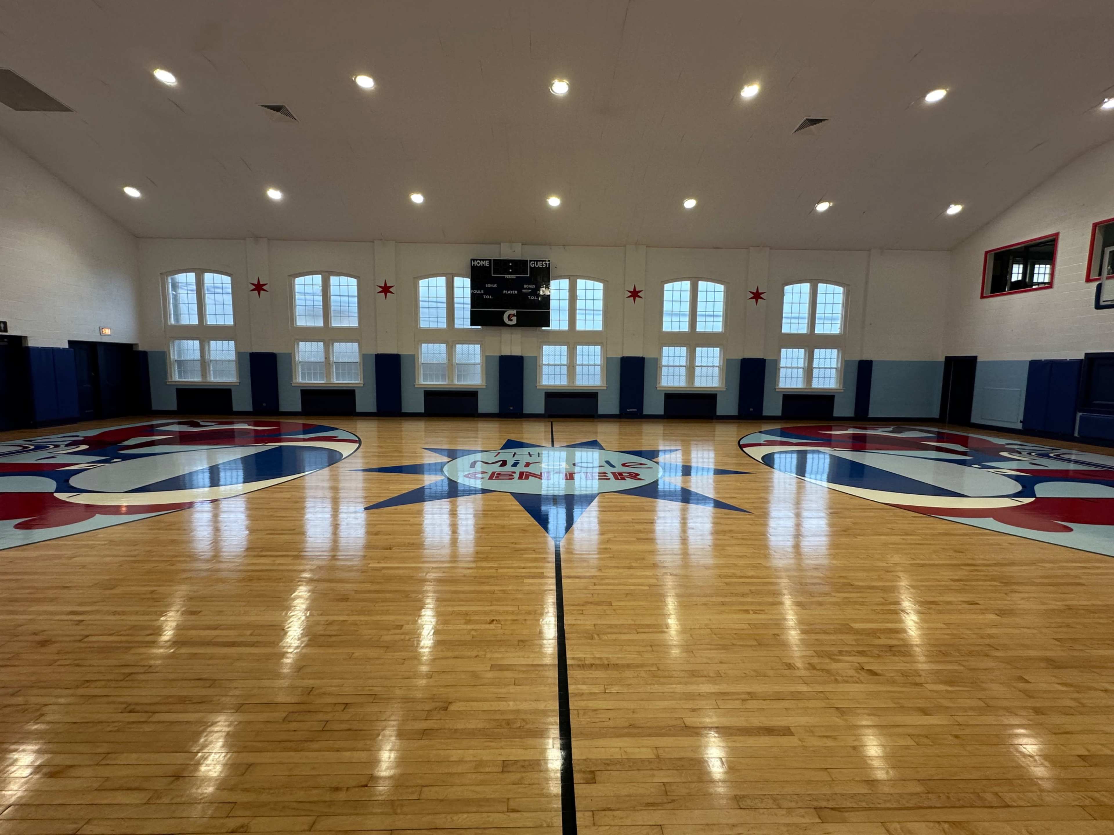 The image shows a basketball gymnasium with a polished wooden floor and a large logo at the center.