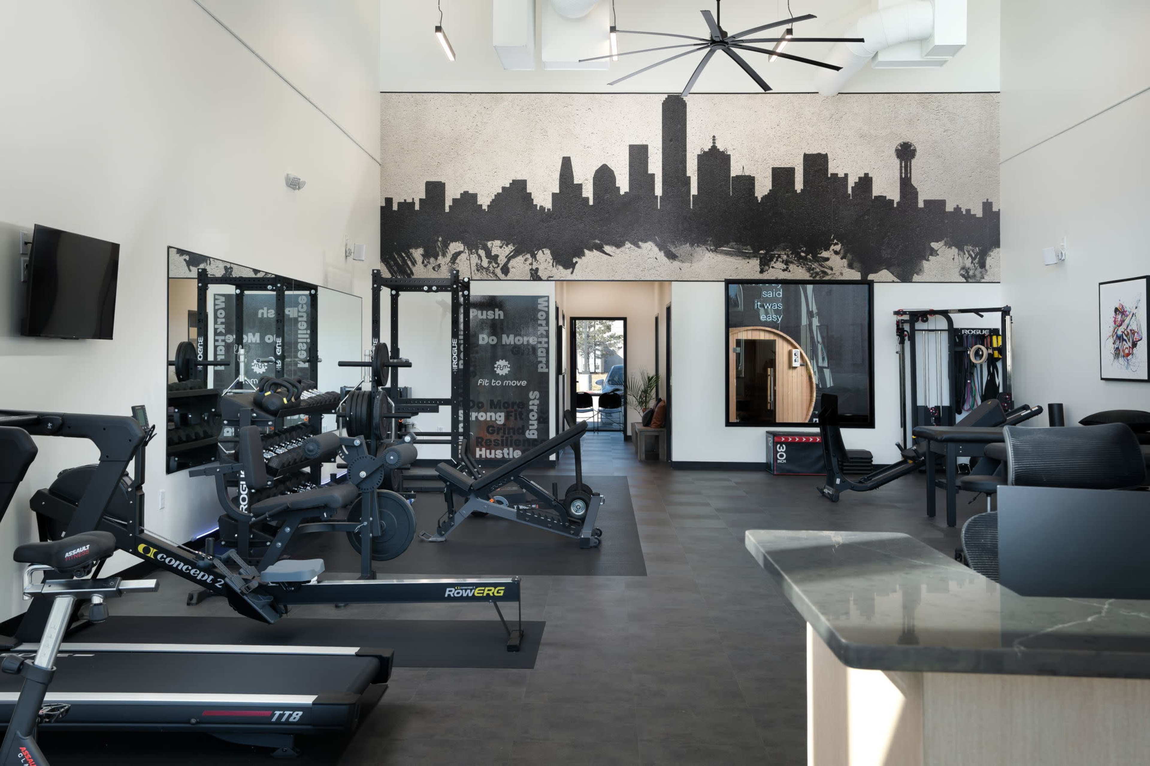 The image shows a modern gym interior featuring fitness equipment, large mirrors, and a mural of a city skyline on the wall.