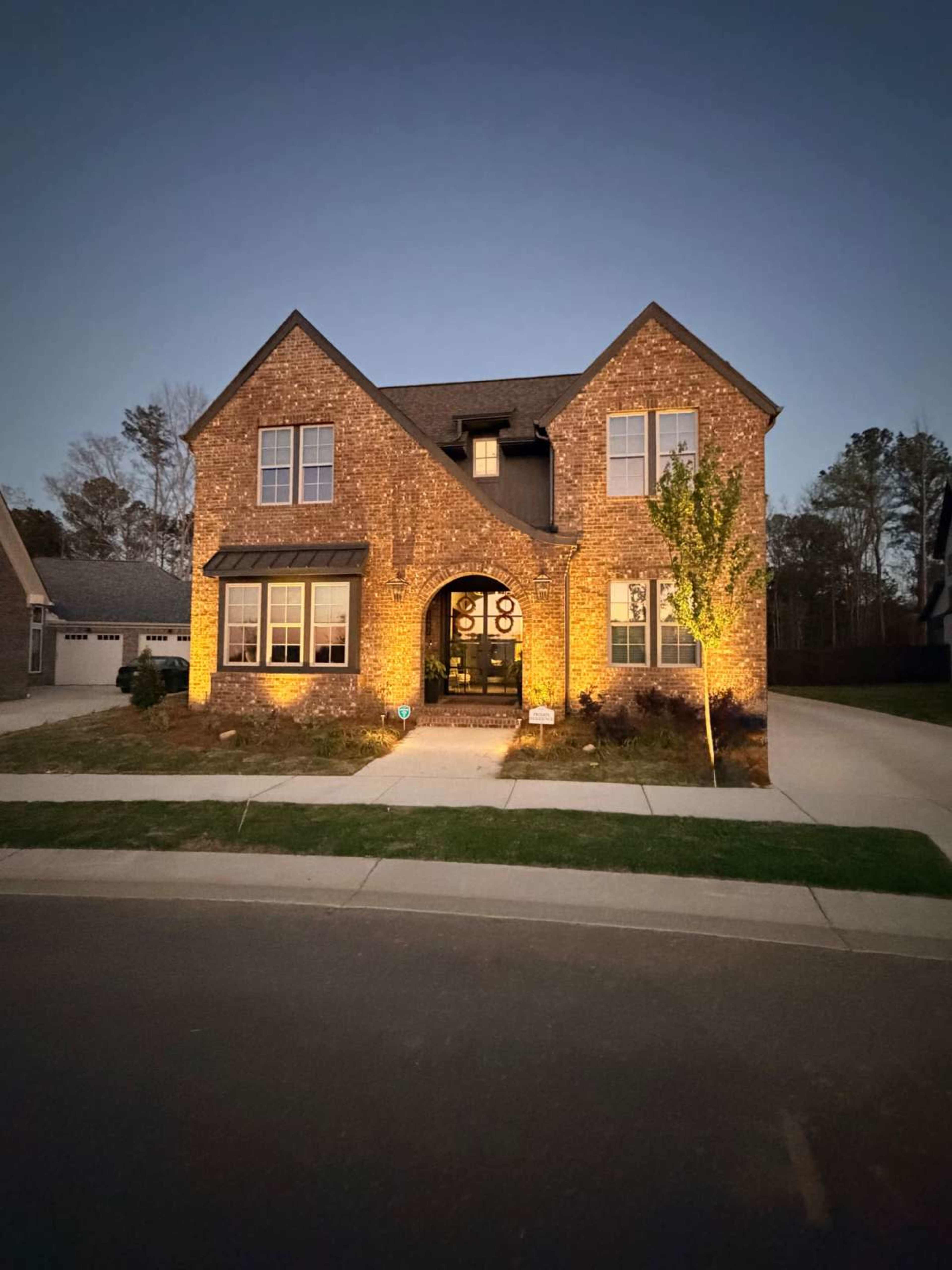 A two-story brick house is illuminated with warm lights, showcasing its front entrance and landscaping during twilight.