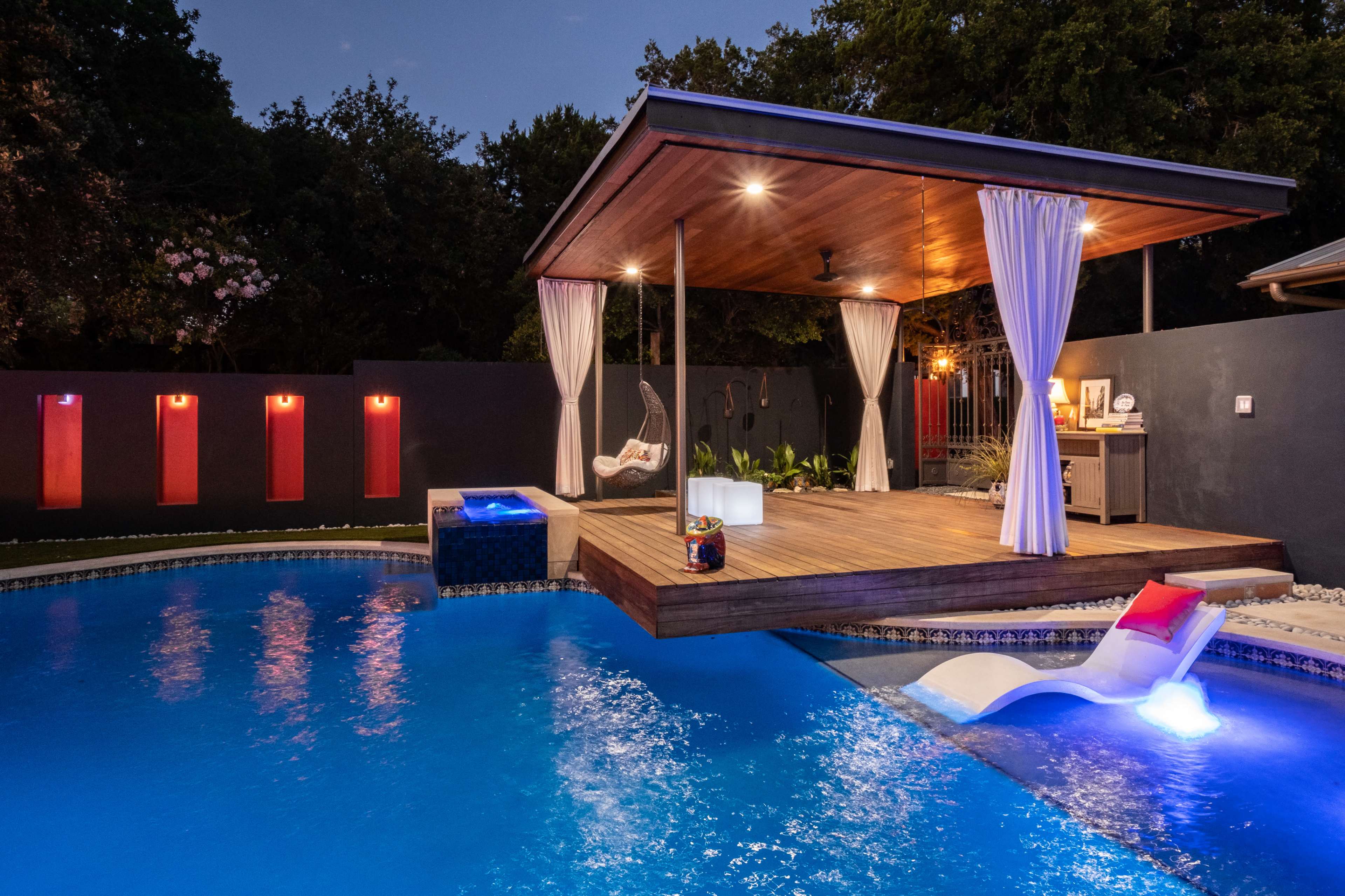 A wooden cabana with draped curtains overlooks a lit pool featuring a spa area, surrounded by trees and a dark wall with vertical lights.