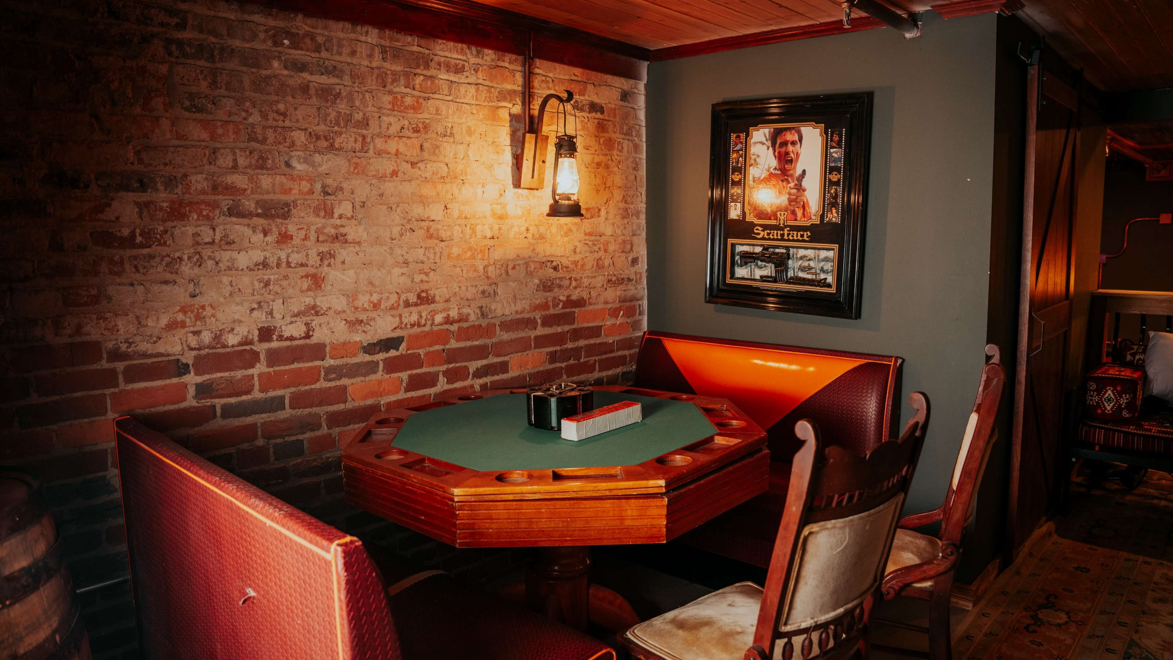 A small, dimly lit room features a green-felt poker table surrounded by upholstered chairs, with exposed brick walls and a Scarface poster hanging nearby.