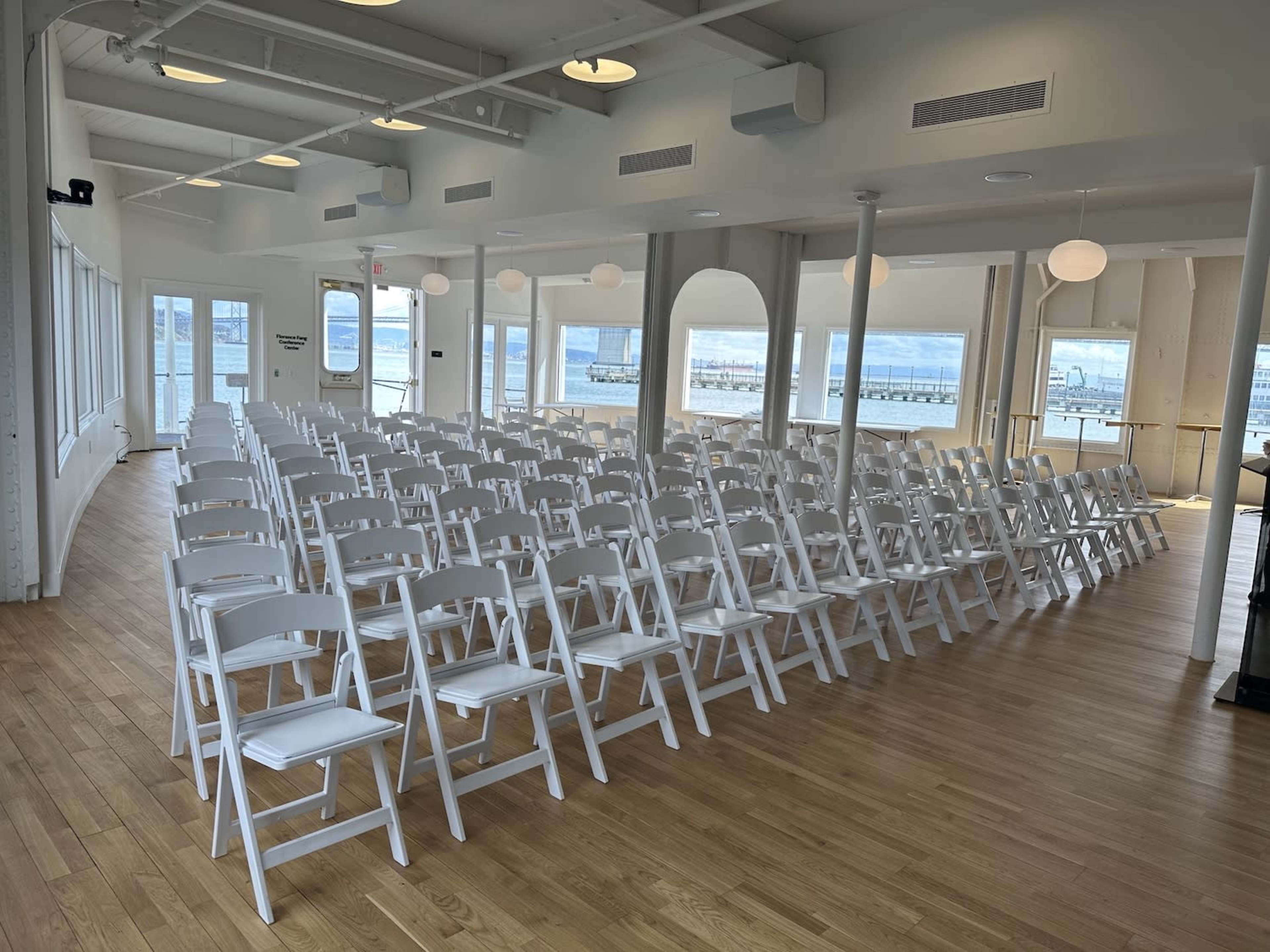 A spacious room set up with rows of white folding chairs arranged in preparation for an event, with large windows providing a view of a body of water.