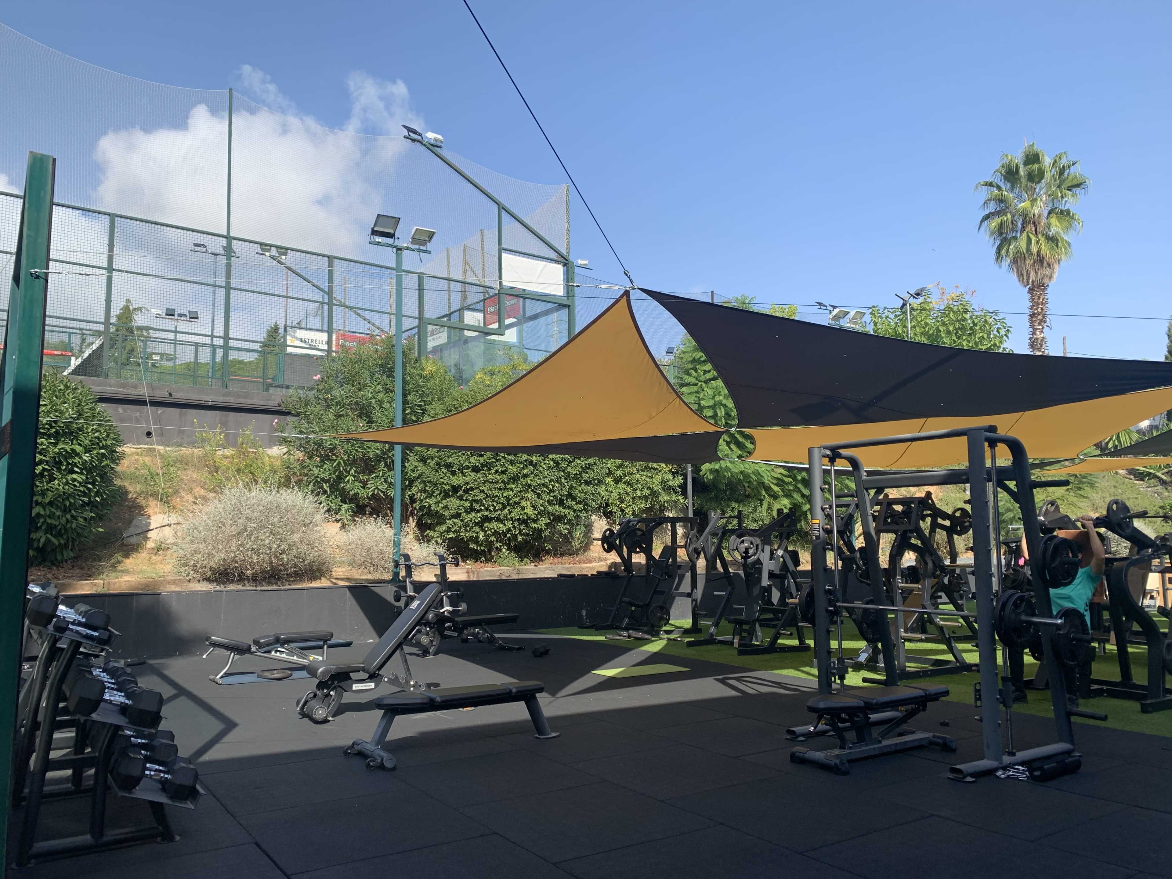 An outdoor gym area with various weightlifting equipment under a shaded canopy and a sports court in the background.
