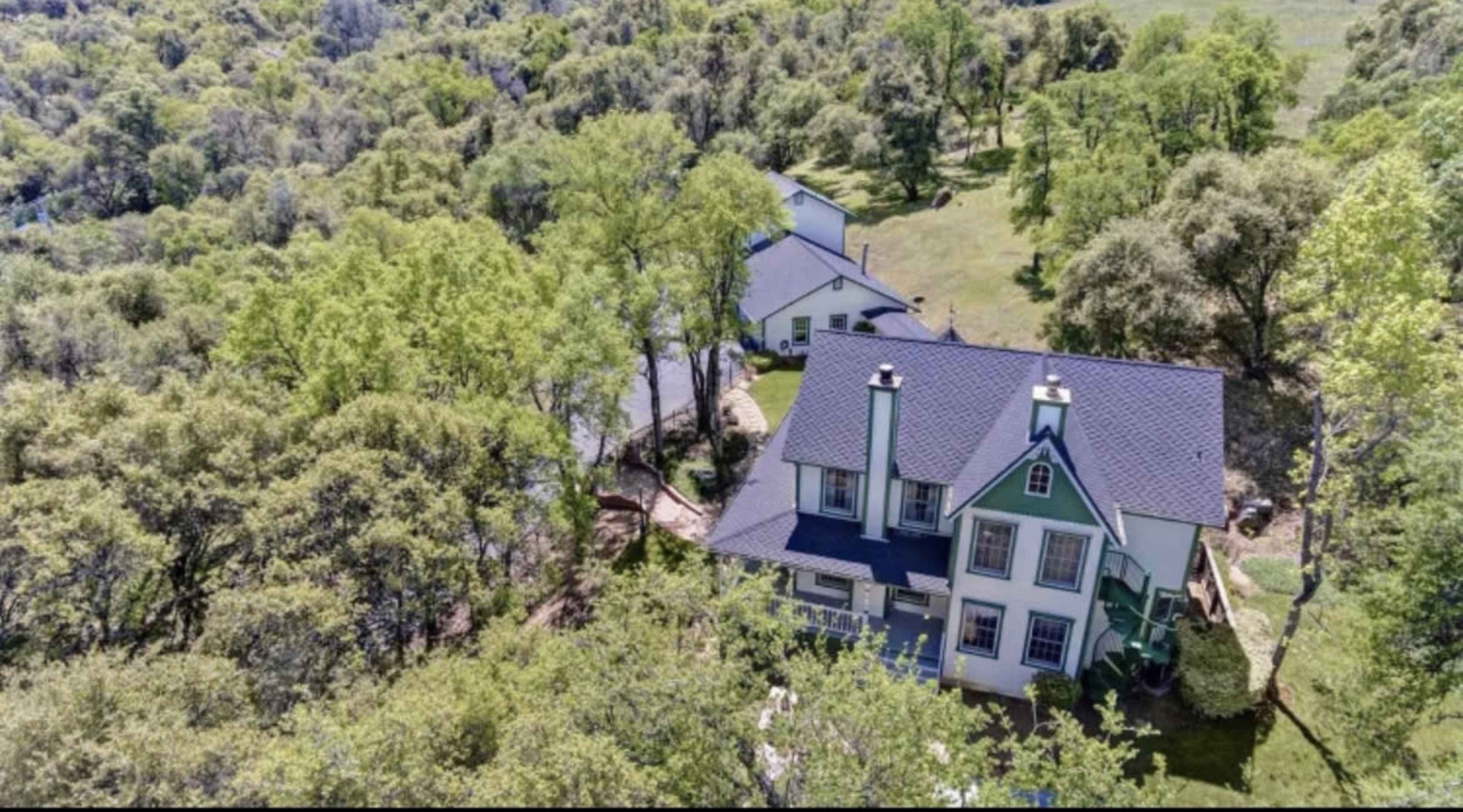 The image shows a large, two-story house surrounded by lush greenery and trees.