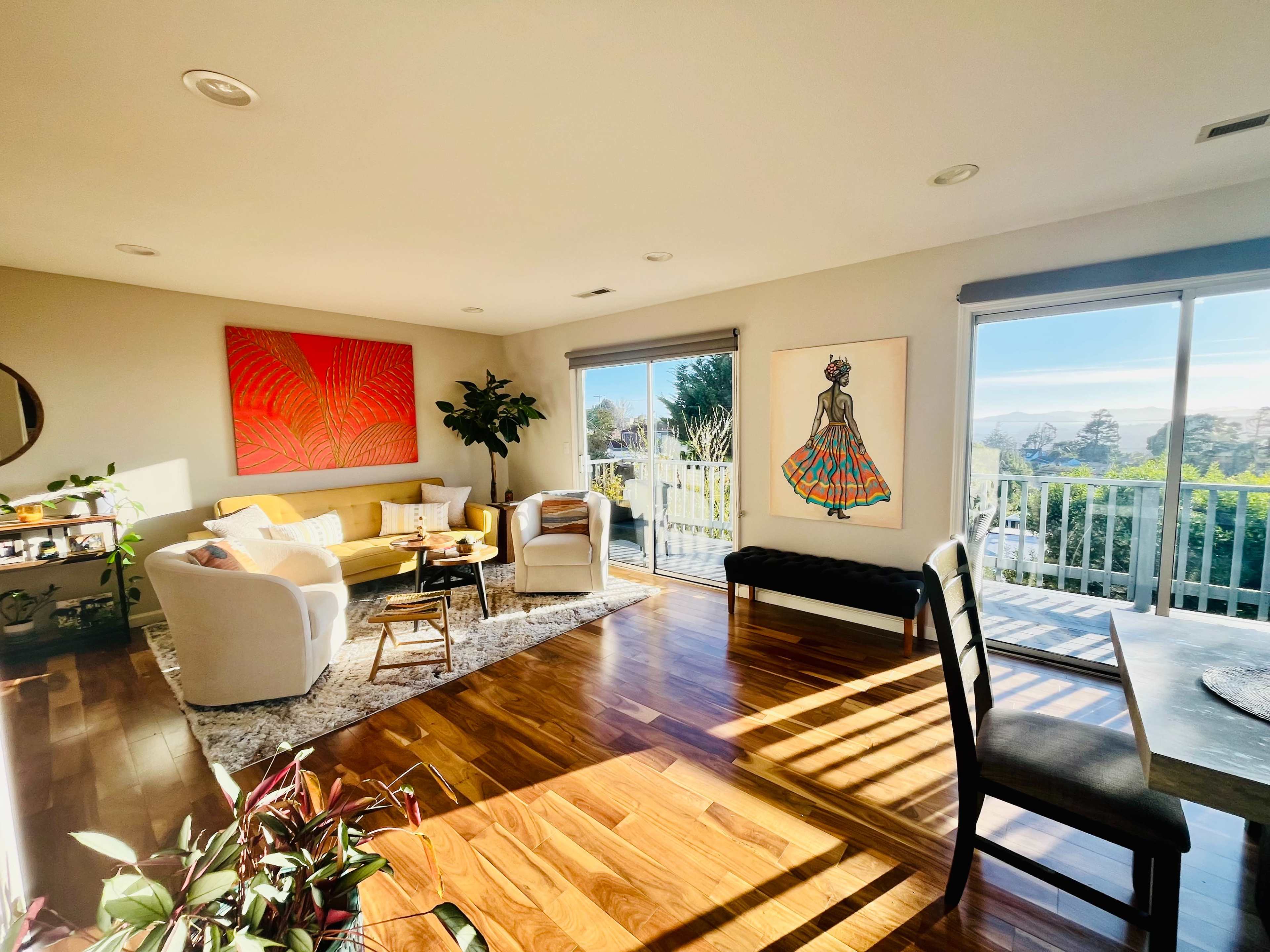 A bright living room features a yellow sofa, two white chairs, a large artwork, and sliding glass doors that open to a balcony with a view.
