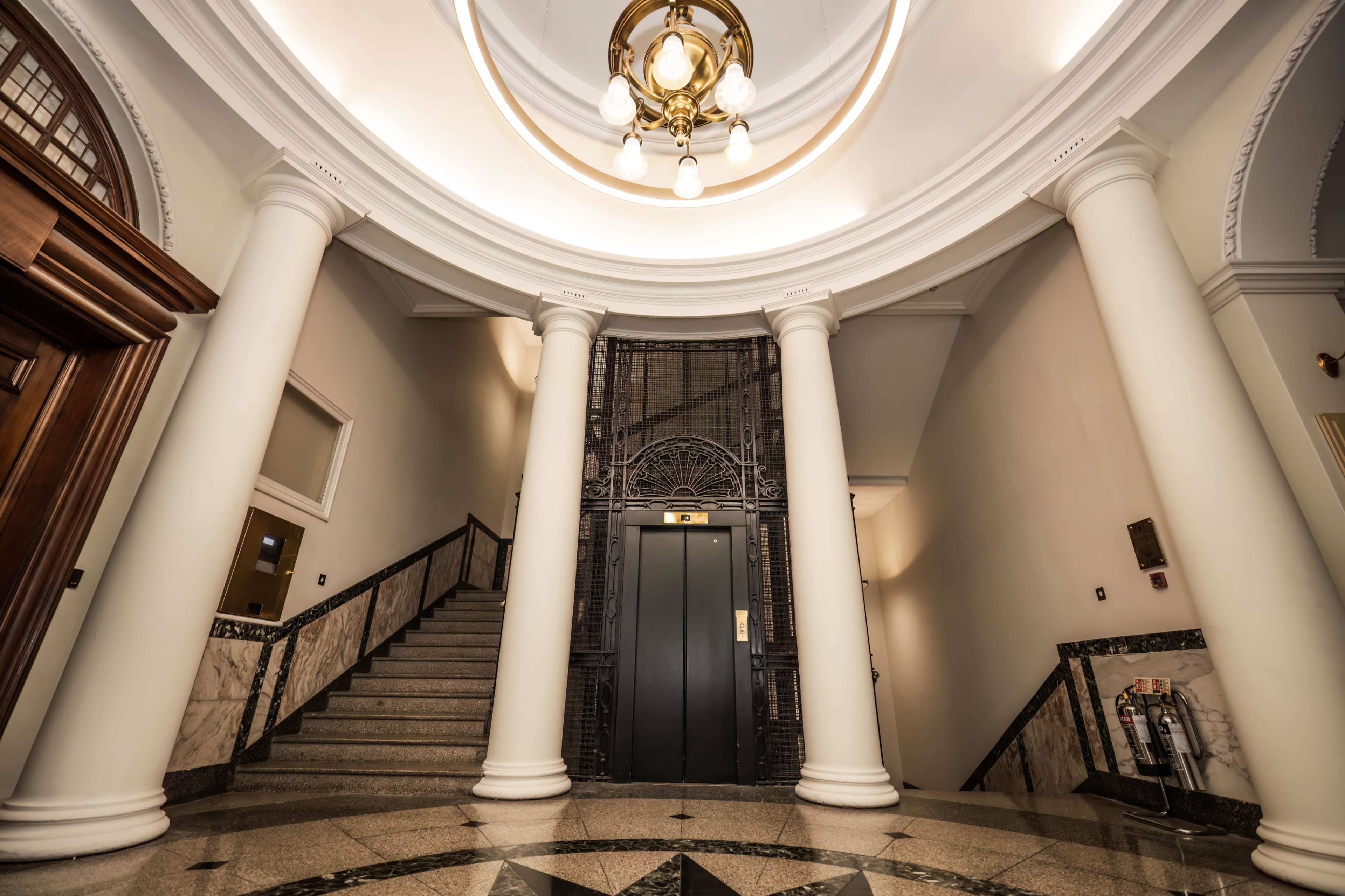 A spacious hallway with elegant columns, a staircase on the left, and an elevator at the center beneath a decorative ceiling light.