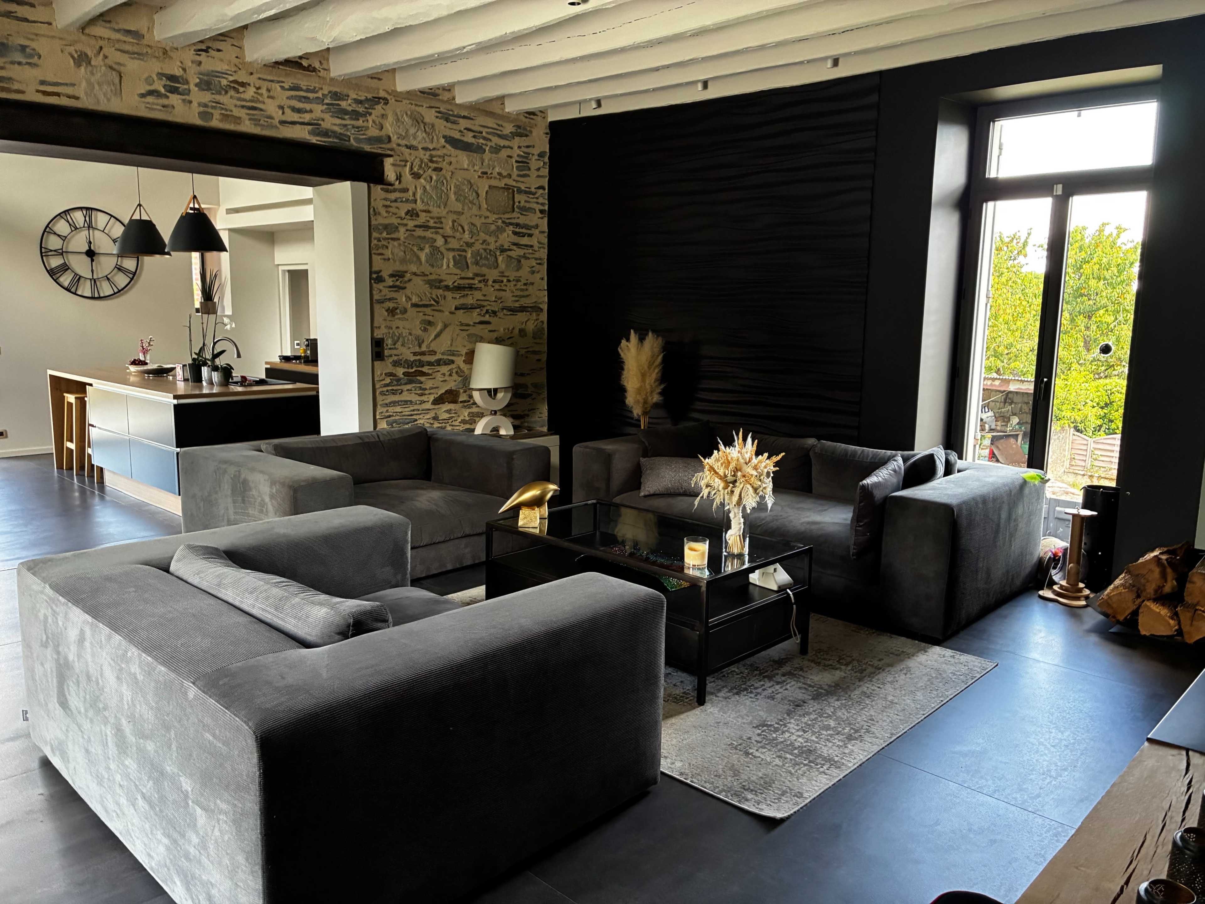 The image shows a modern living room with two gray sofas, a glass coffee table, and a stone wall, with an open view to a kitchen in the background.