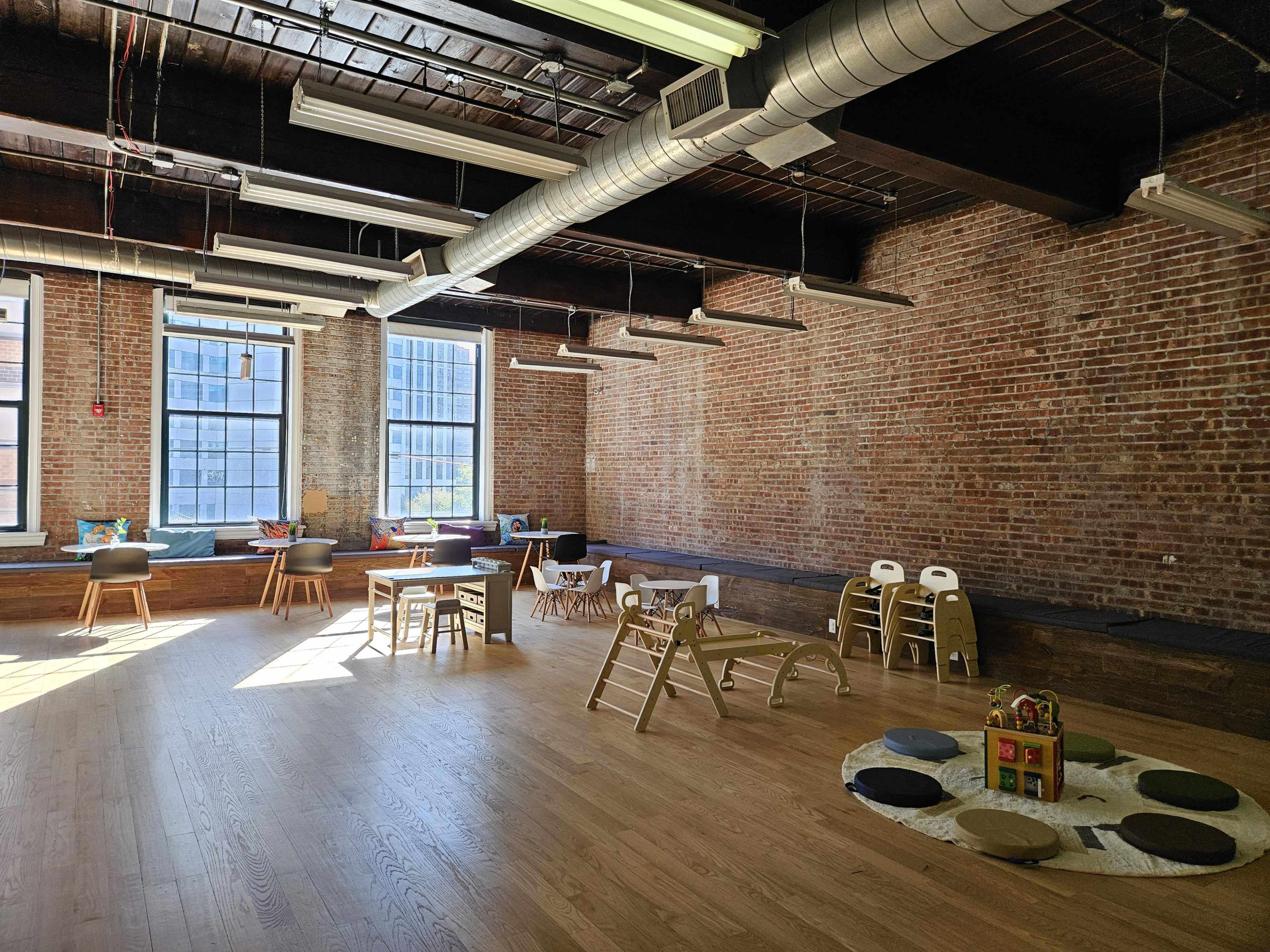 The image shows a spacious room with exposed brick walls, wooden flooring, and various play equipment arranged for children.