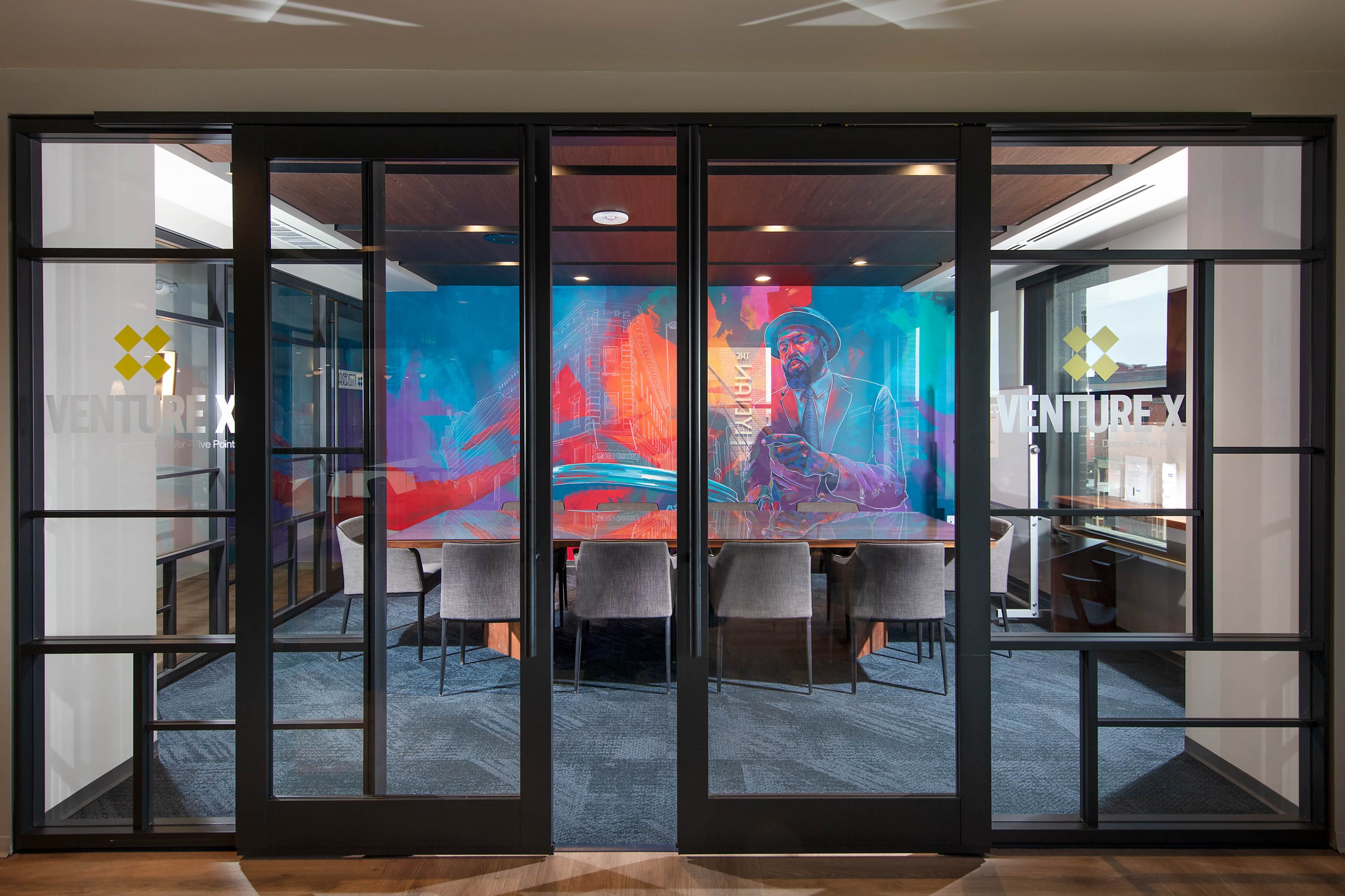 A modern conference room with glass doors, featuring a colorful mural and a long table surrounded by chairs.