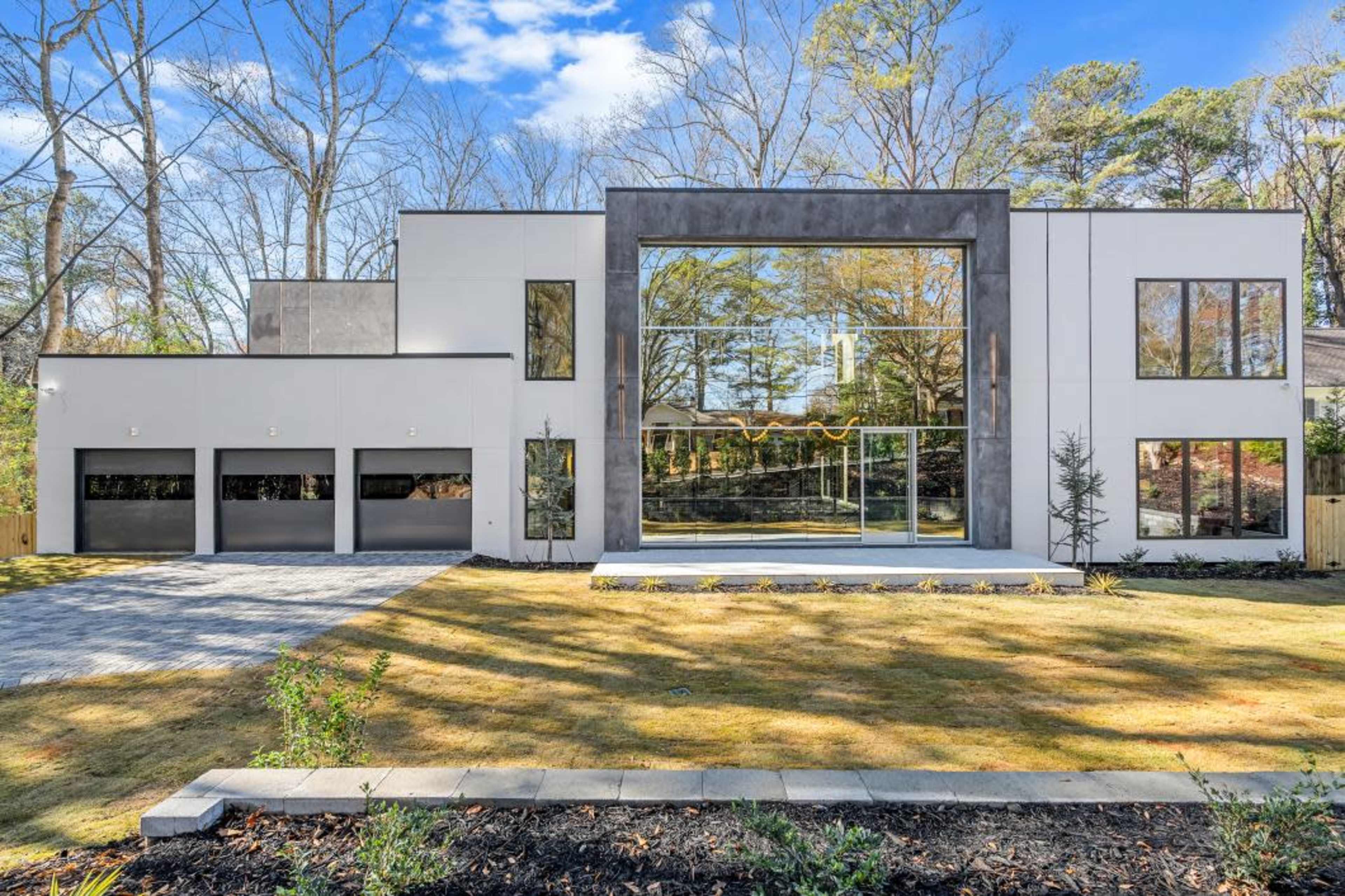 A modern two-story house with large glass windows and a landscaped front yard is set against a backdrop of trees.
