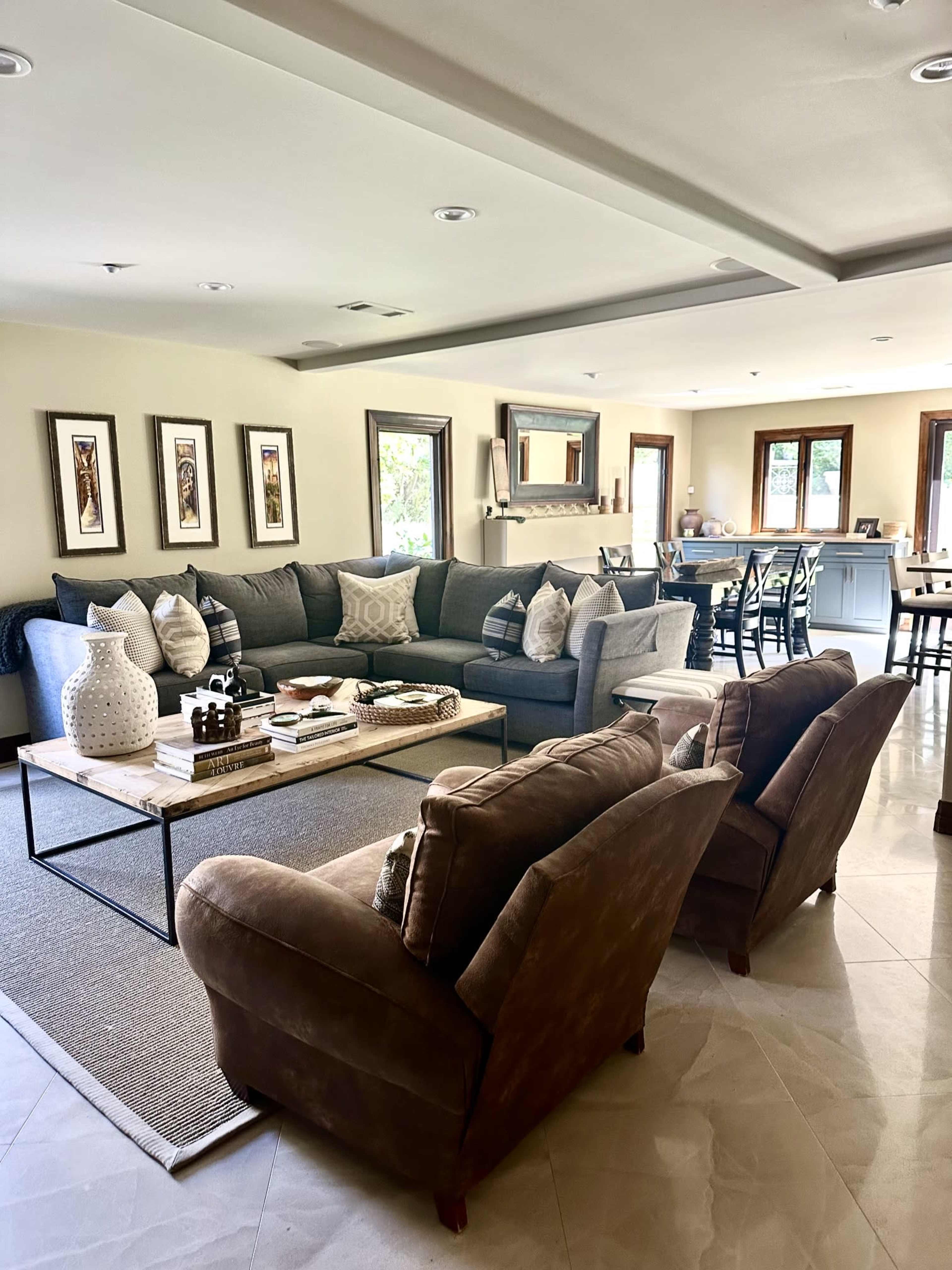 A spacious living room features a gray sectional sofa with decorative pillows, a coffee table in the center, and two brown chairs, along with framed artwork on the walls and a view of a dining area in the background.