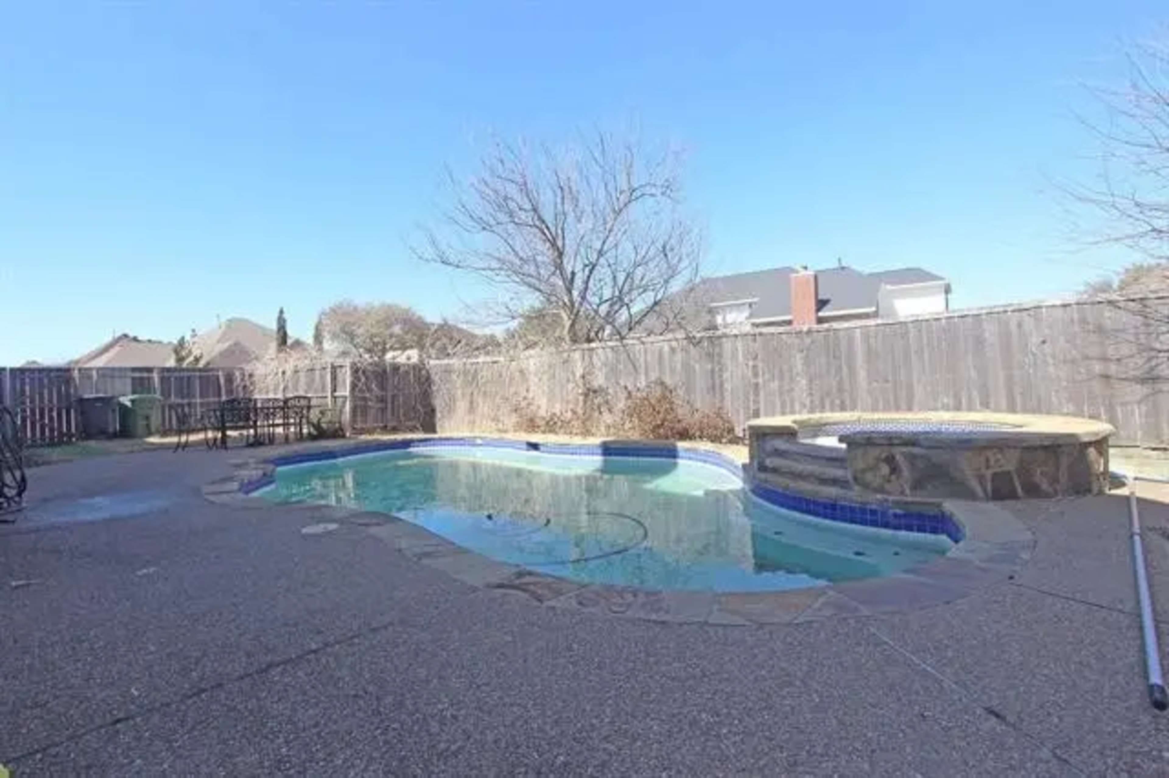 A partially empty swimming pool surrounded by a concrete patio and a wooden fence.