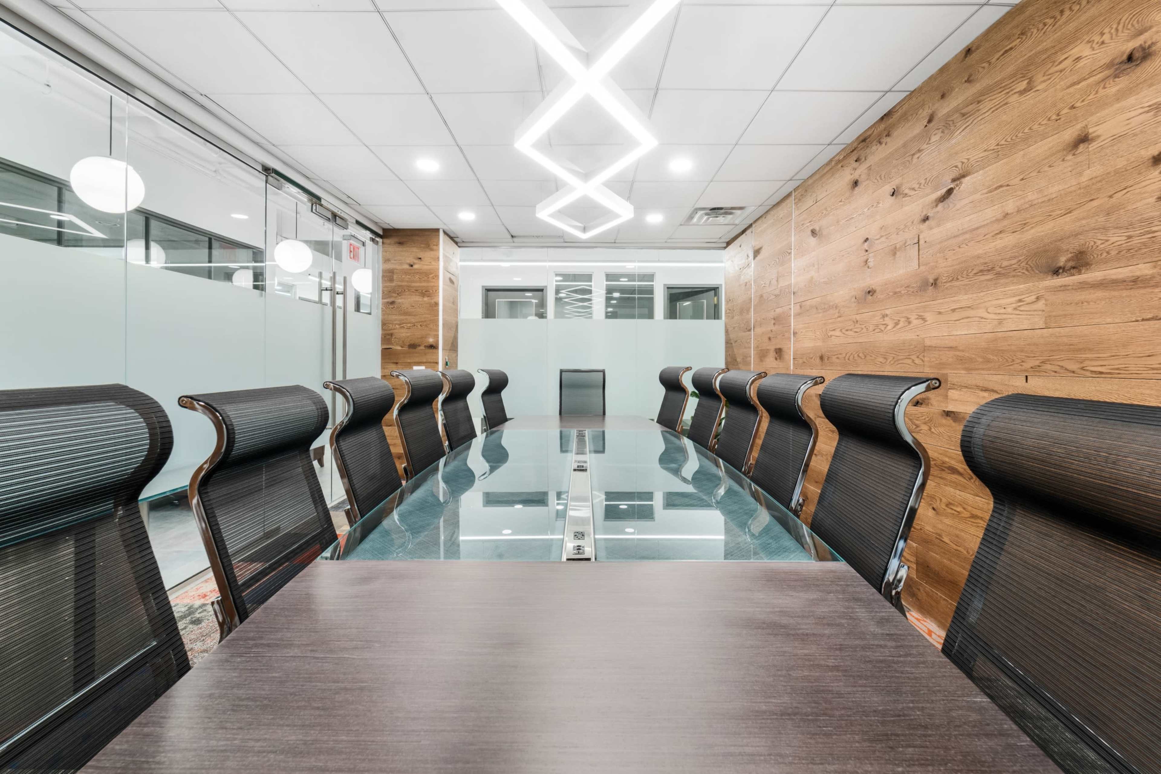 A long conference table with black chairs is in a modern meeting room featuring wooden walls and a geometric light fixture above.