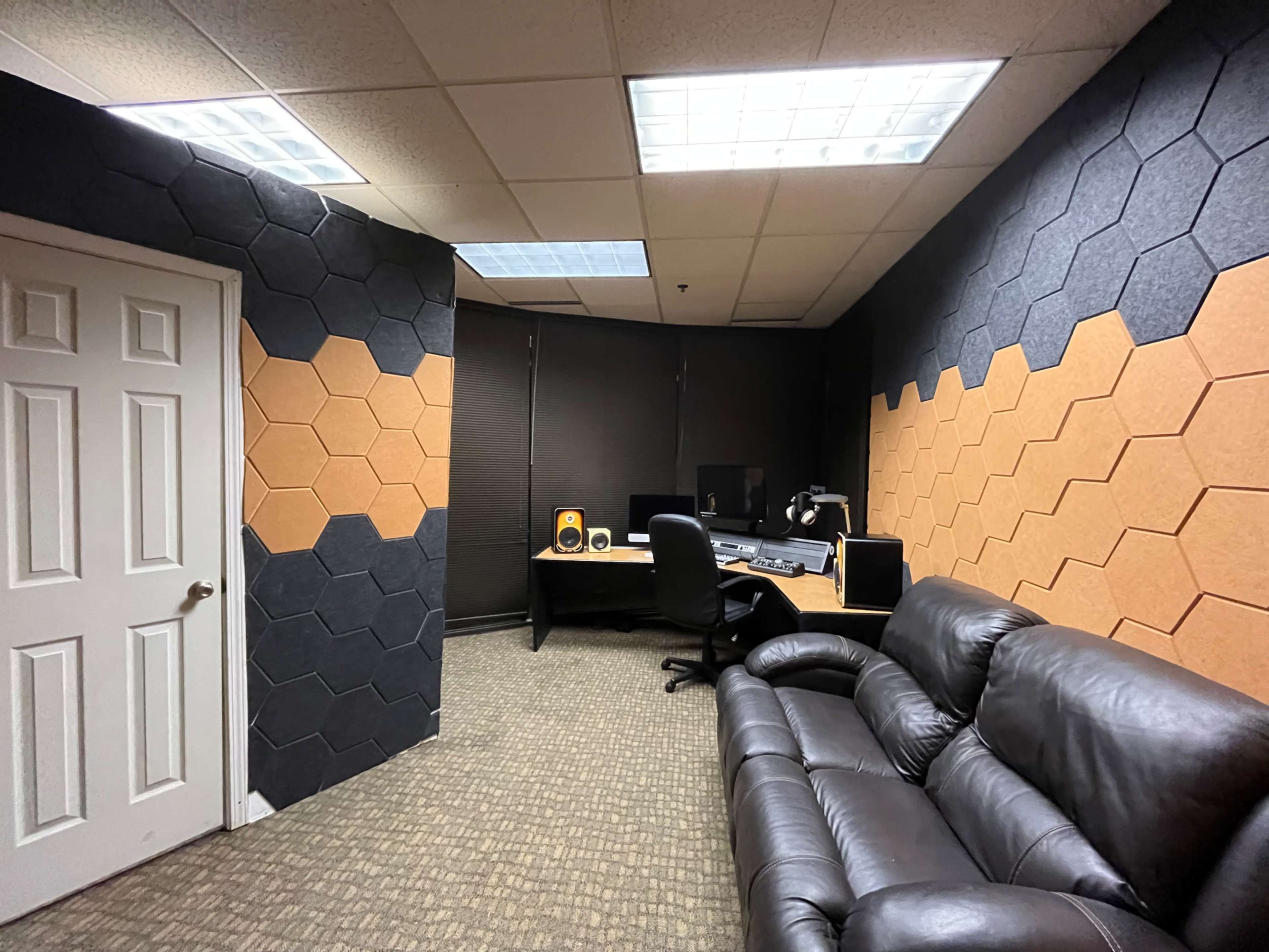 A small recording studio featuring hexagon-patterned soundproofing panels on the walls, a desk with computer equipment, and a leather sofa.