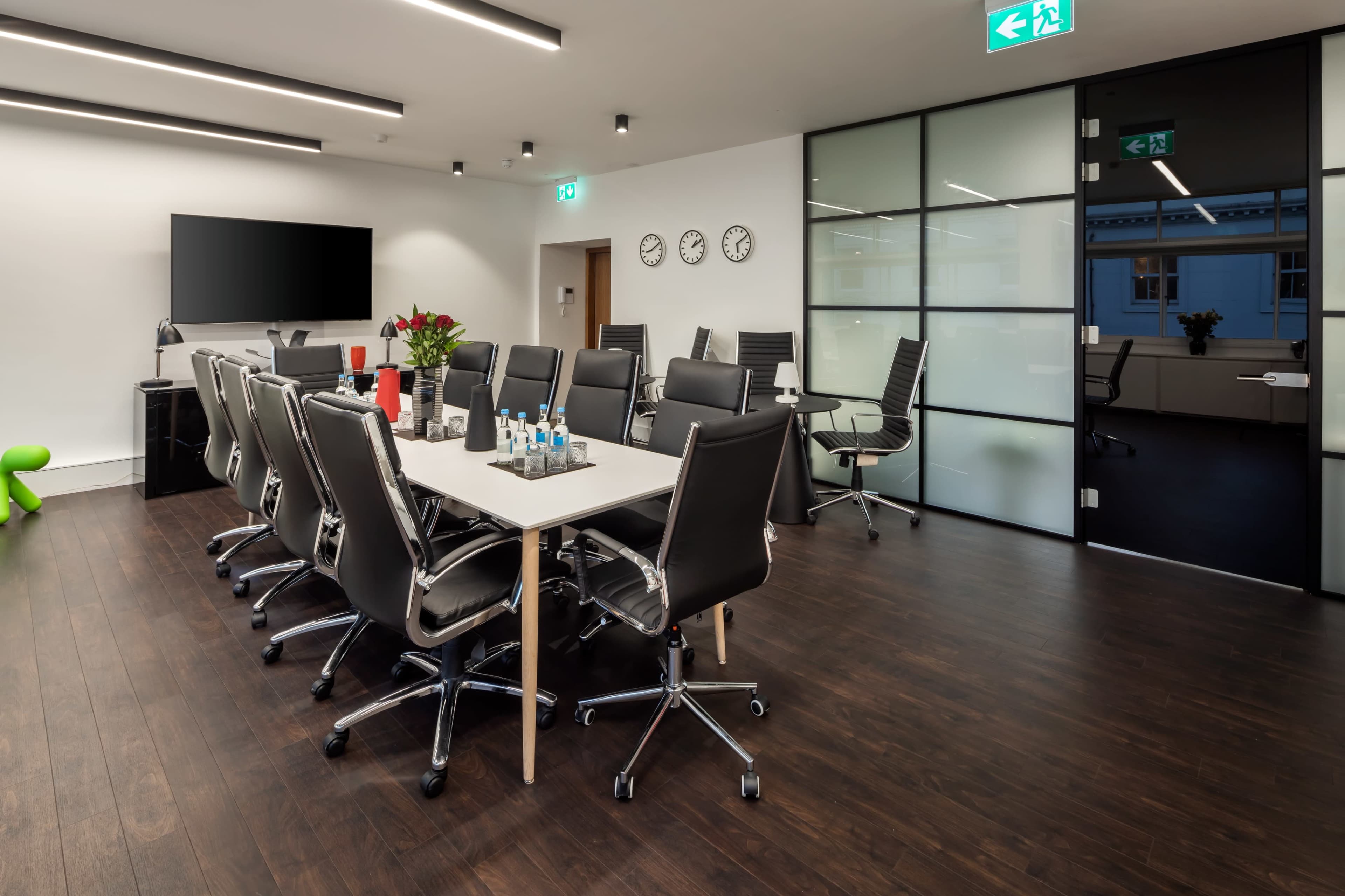 A modern conference room features a long table surrounded by black office chairs, with a large screen on one wall and glass doors.