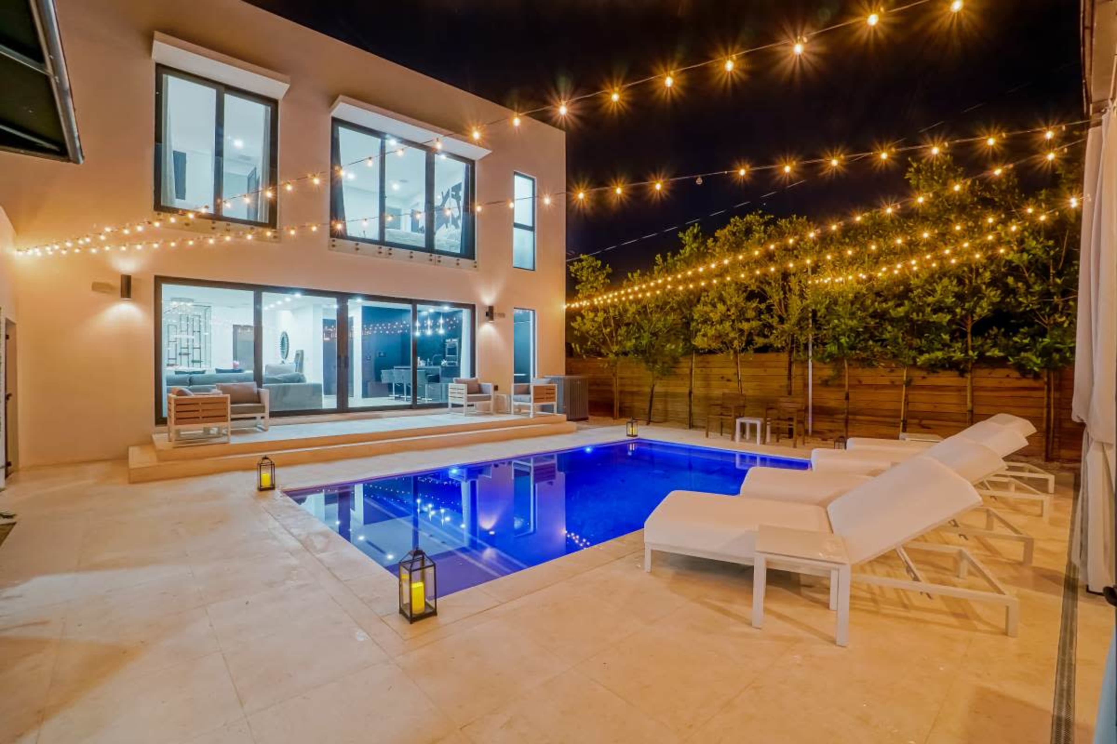 The image shows a modern outdoor pool area with several lounge chairs, surrounded by trees and string lights, illuminated at night.