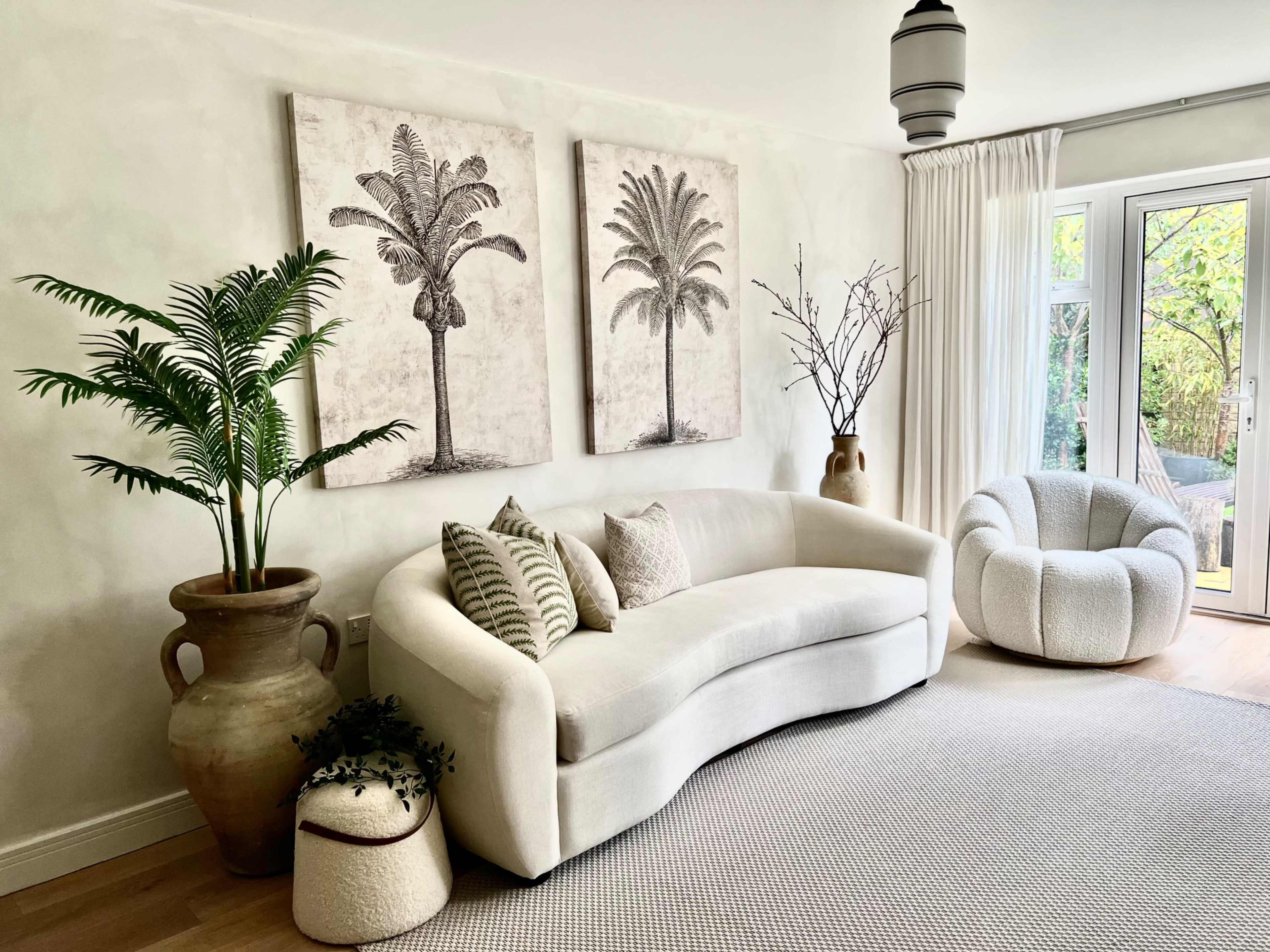 A light-colored living room features a curved sofa, decorative palm prints on the wall, and various plants in stylish pots.