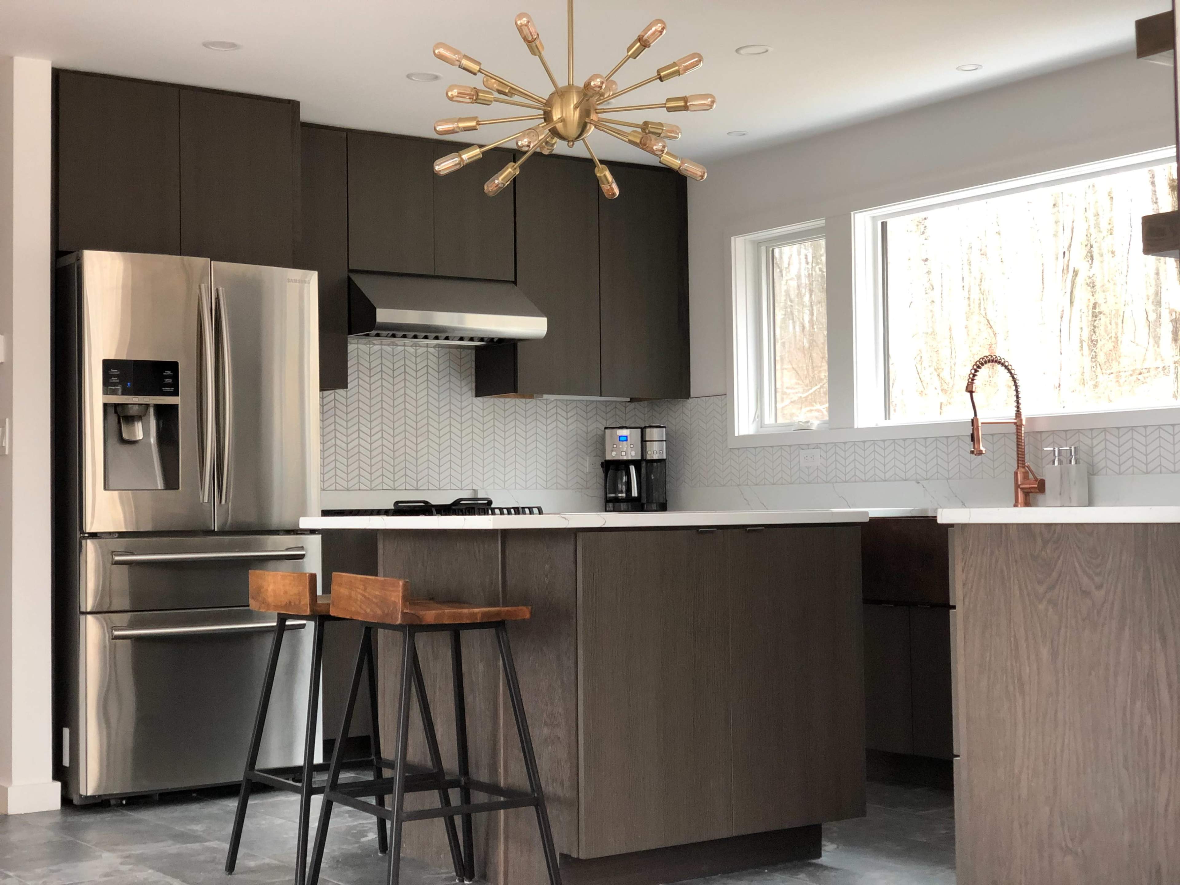 A modern kitchen featuring dark cabinetry, stainless steel appliances, and a geometric backsplash, illuminated by a unique pendant light.