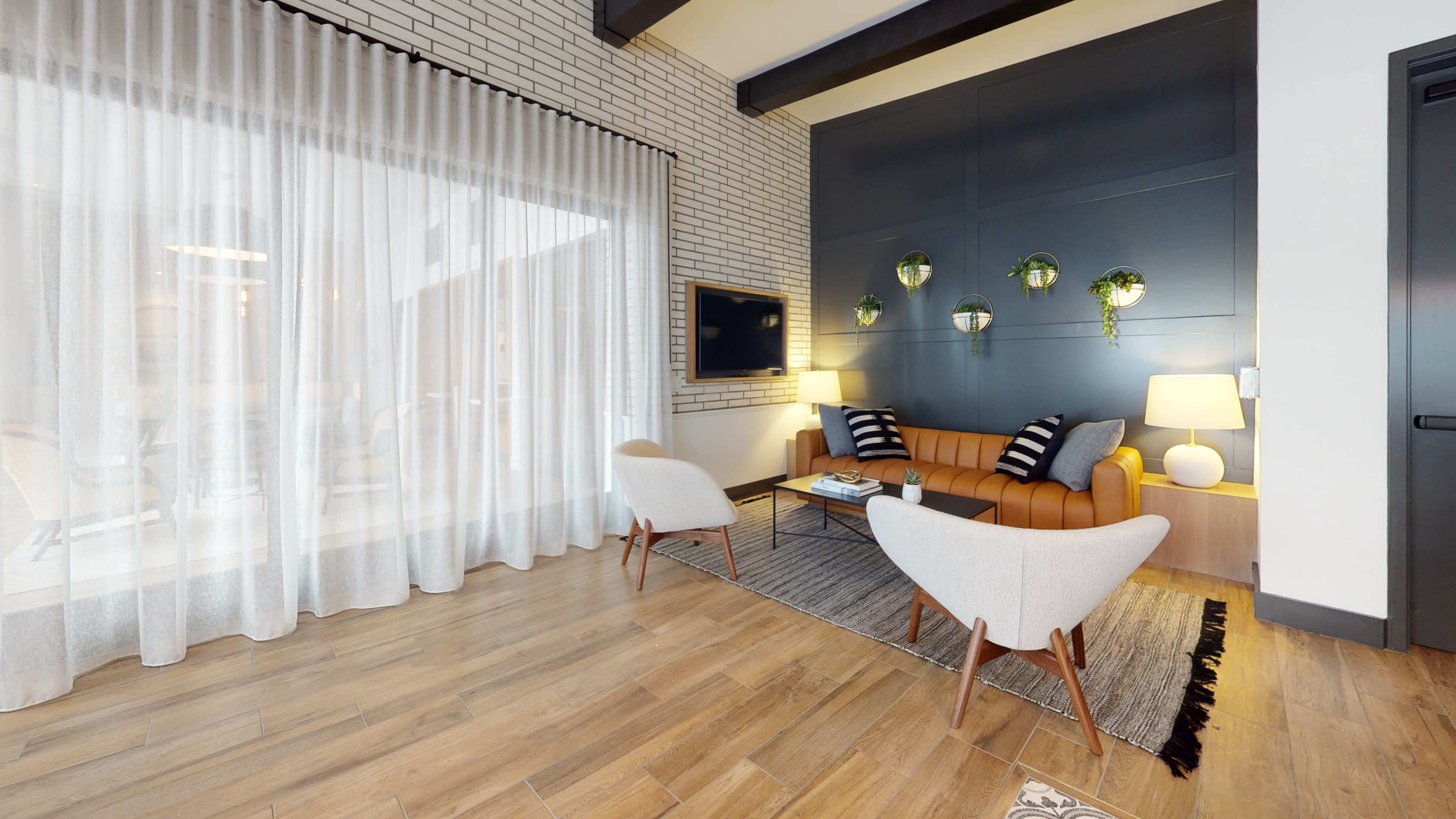 A modern living room features a tan leather couch, two light-colored chairs, and a large window with sheer curtains, accented by a wall-mounted TV and green wall planters.