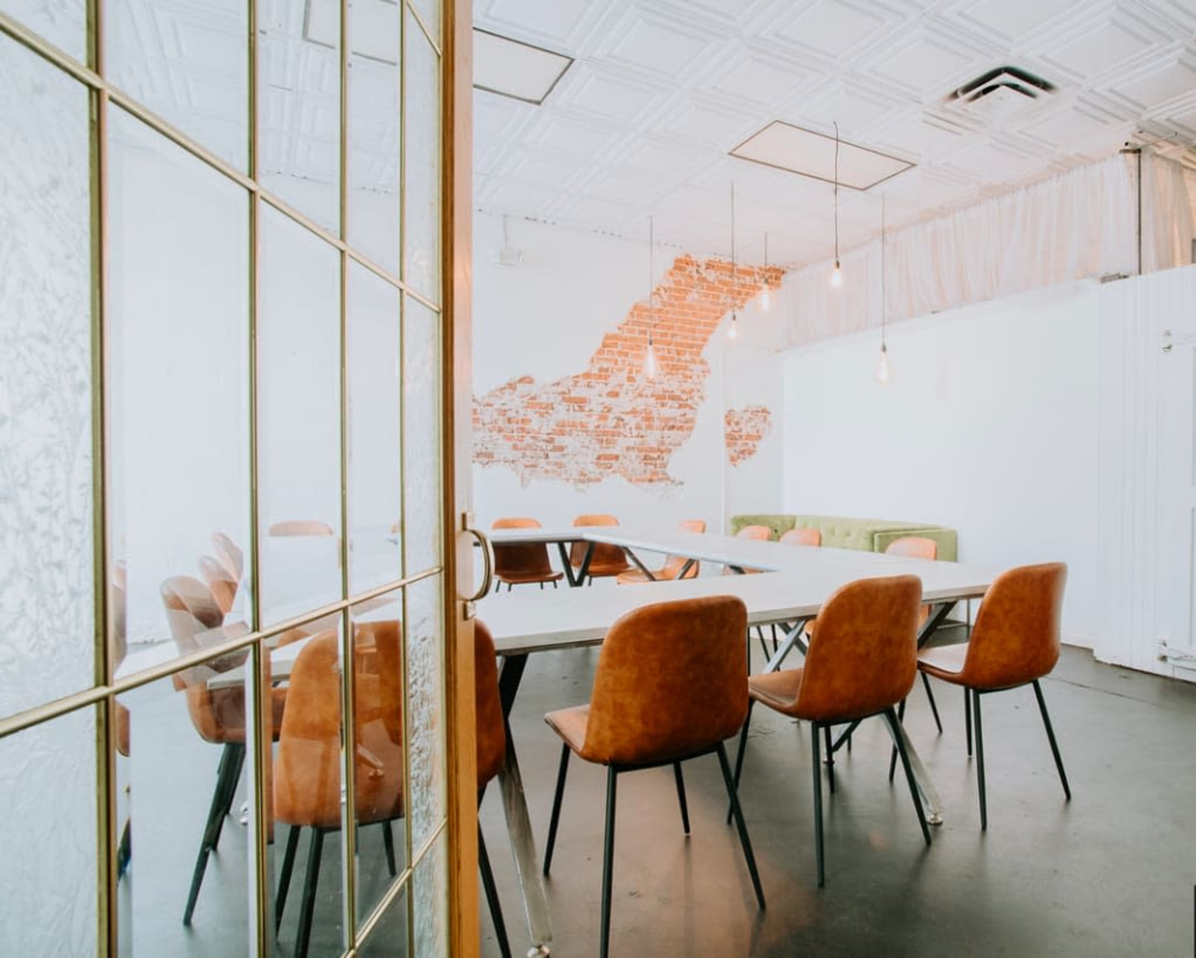 The image shows a modern meeting room with a long table surrounded by brown chairs and a partially exposed brick wall.