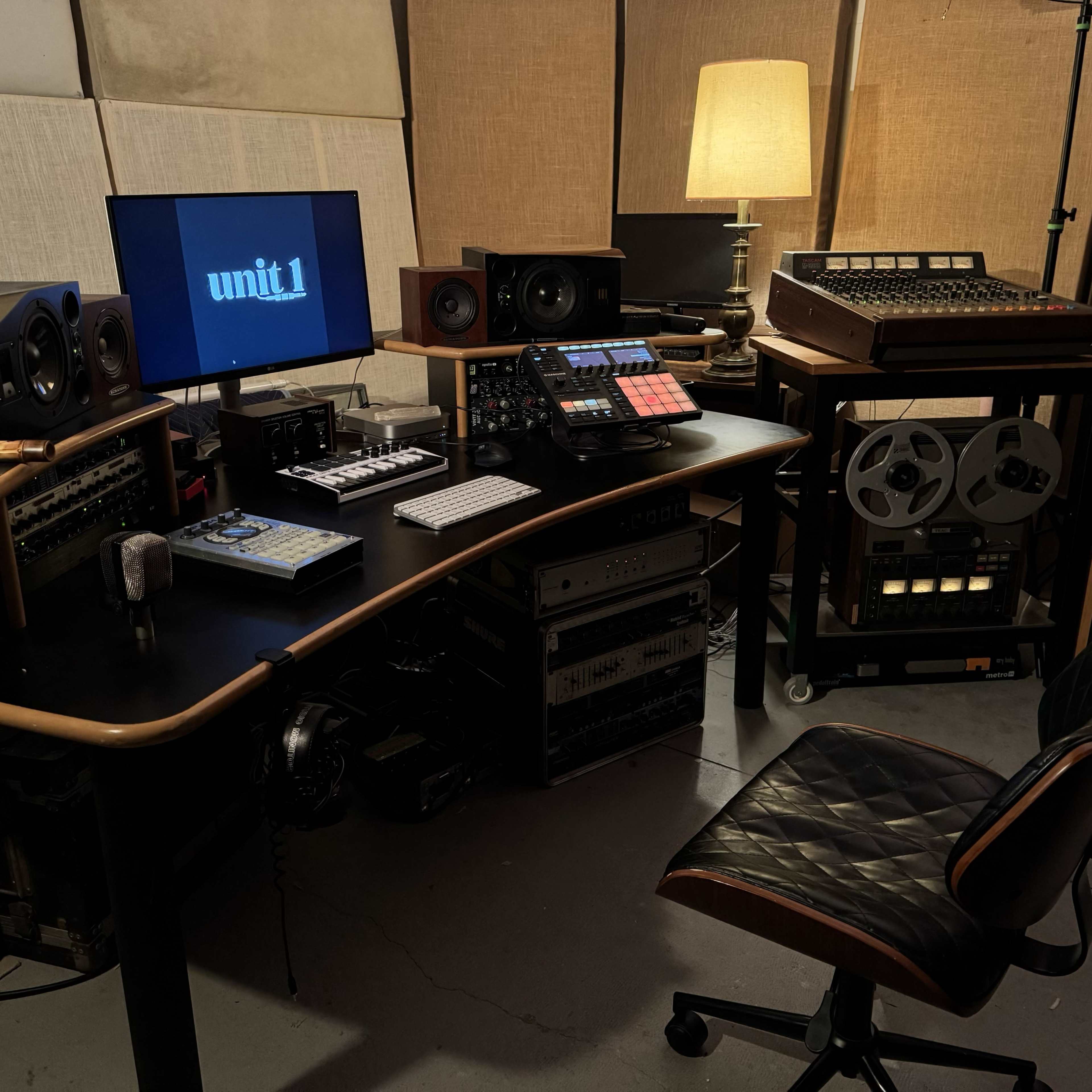A music production workspace features a black desk with audio equipment, a keyboard, a computer monitor displaying "unit1," and surrounding vintage recording devices, illuminated by a lamp.