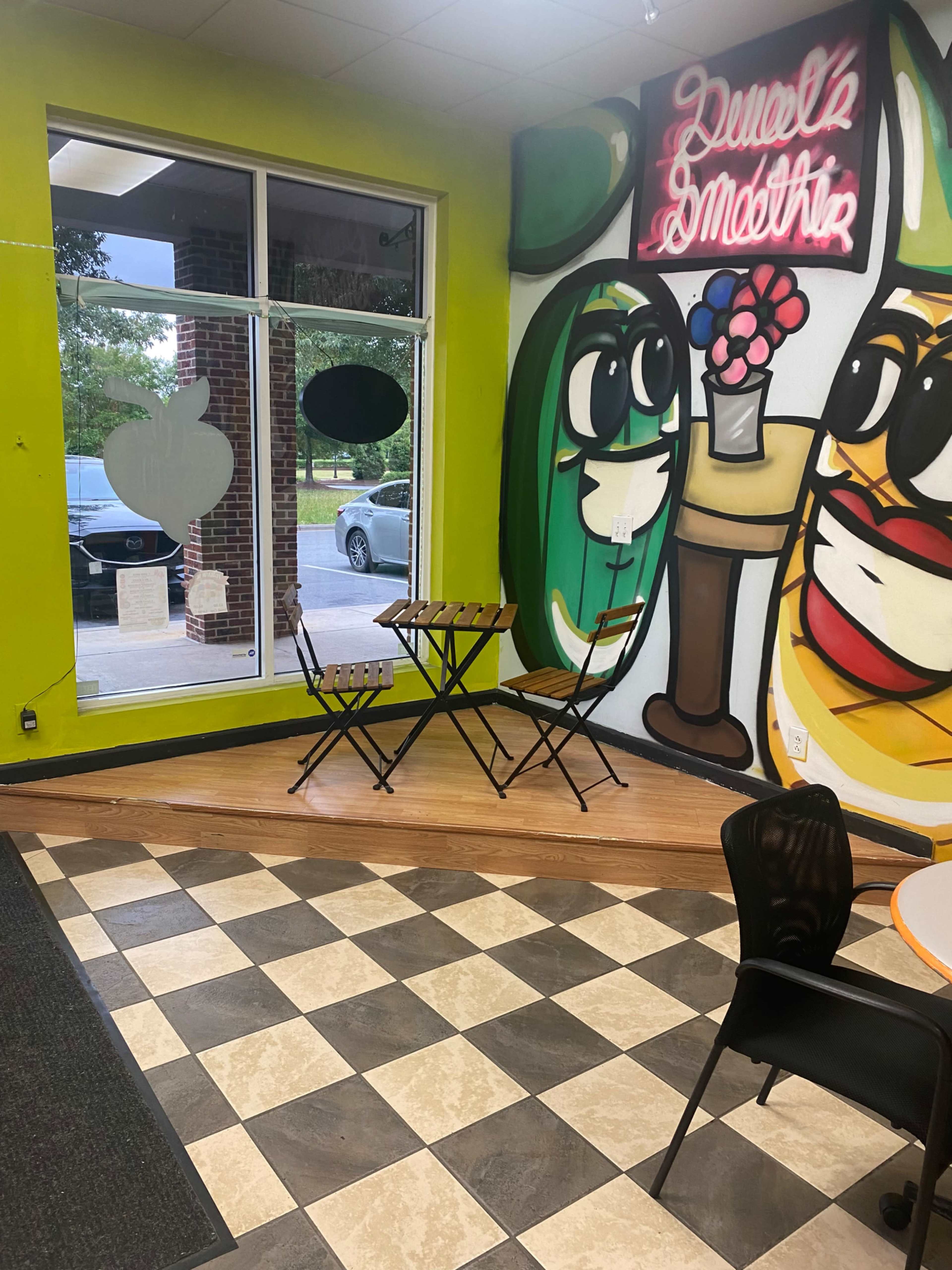 A small café interior with a colorful mural and two metal chairs around a folding table near a large window.