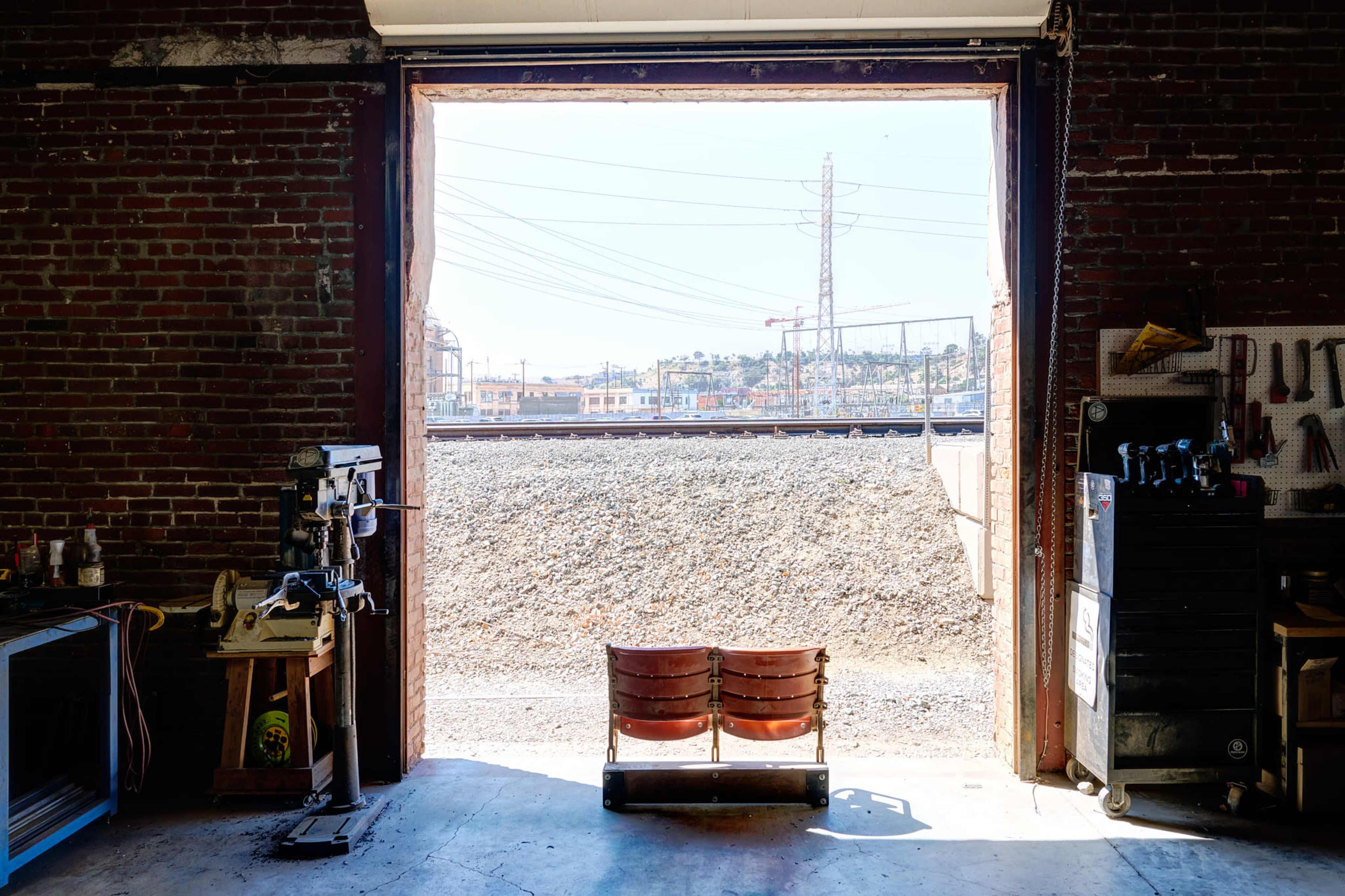 A pair of wooden chairs sits in the open doorway of a workshop, leading to a view of a gravel pathway and distant railroad tracks.