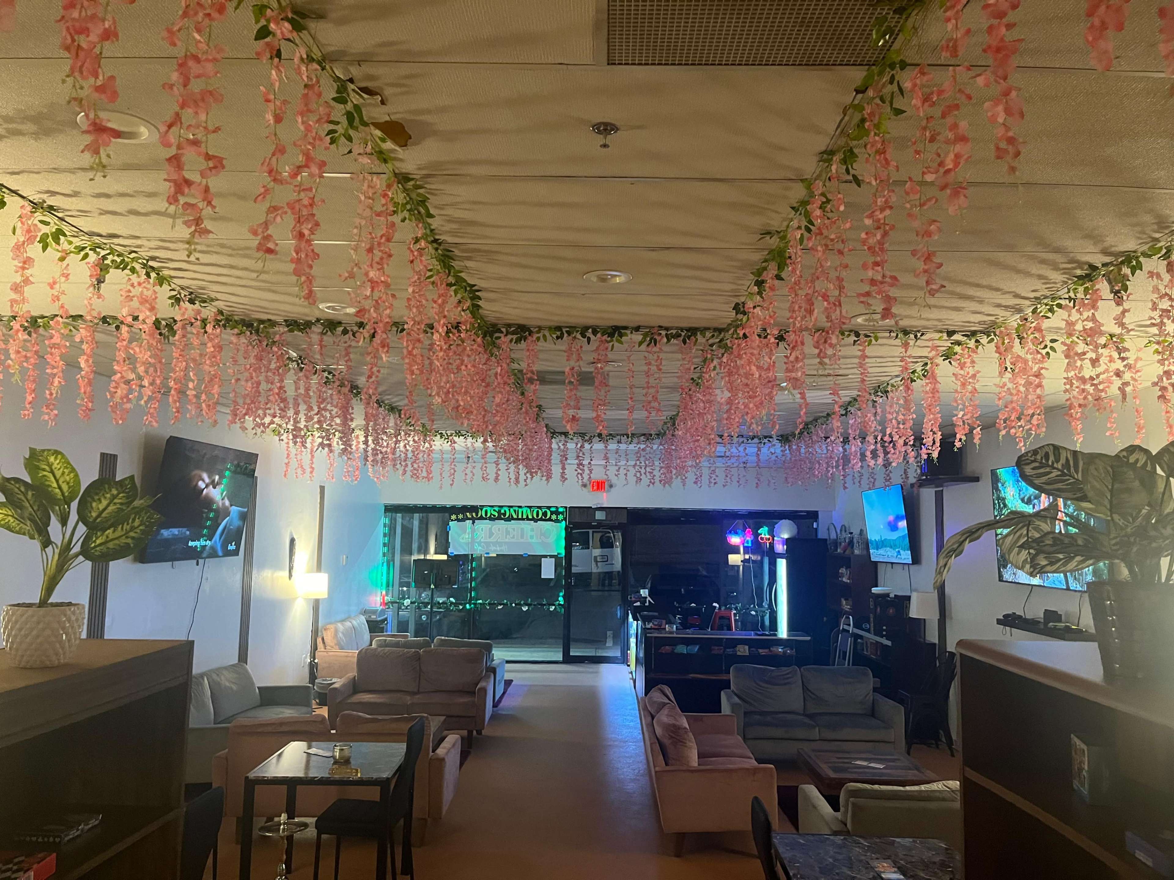The interior of a lounge features pink floral decorations hanging from the ceiling, with various seating areas and multiple televisions displaying content.