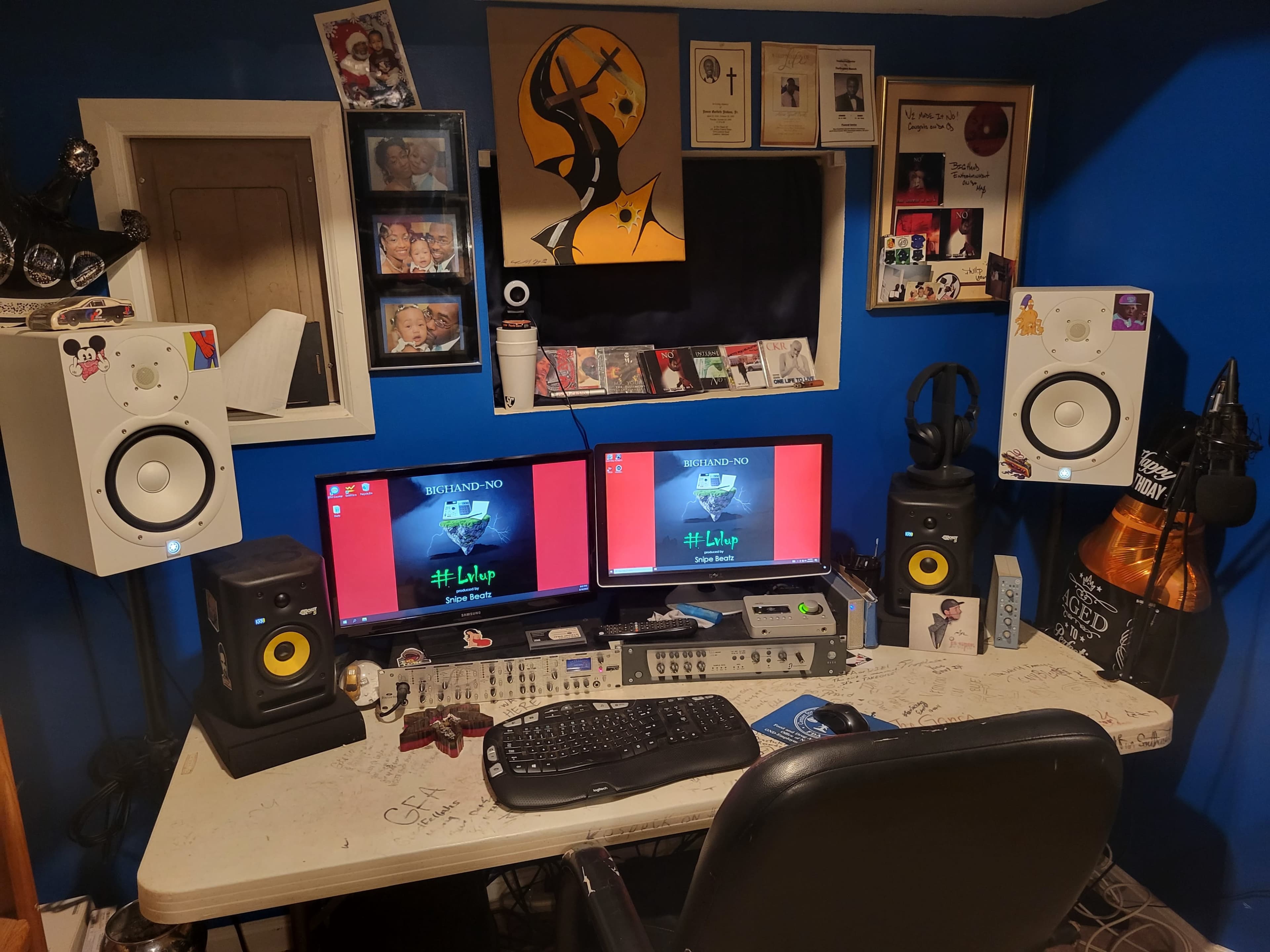 A cluttered workspace features two monitors, audio equipment, speakers, and numerous awards and photos on a blue wall.