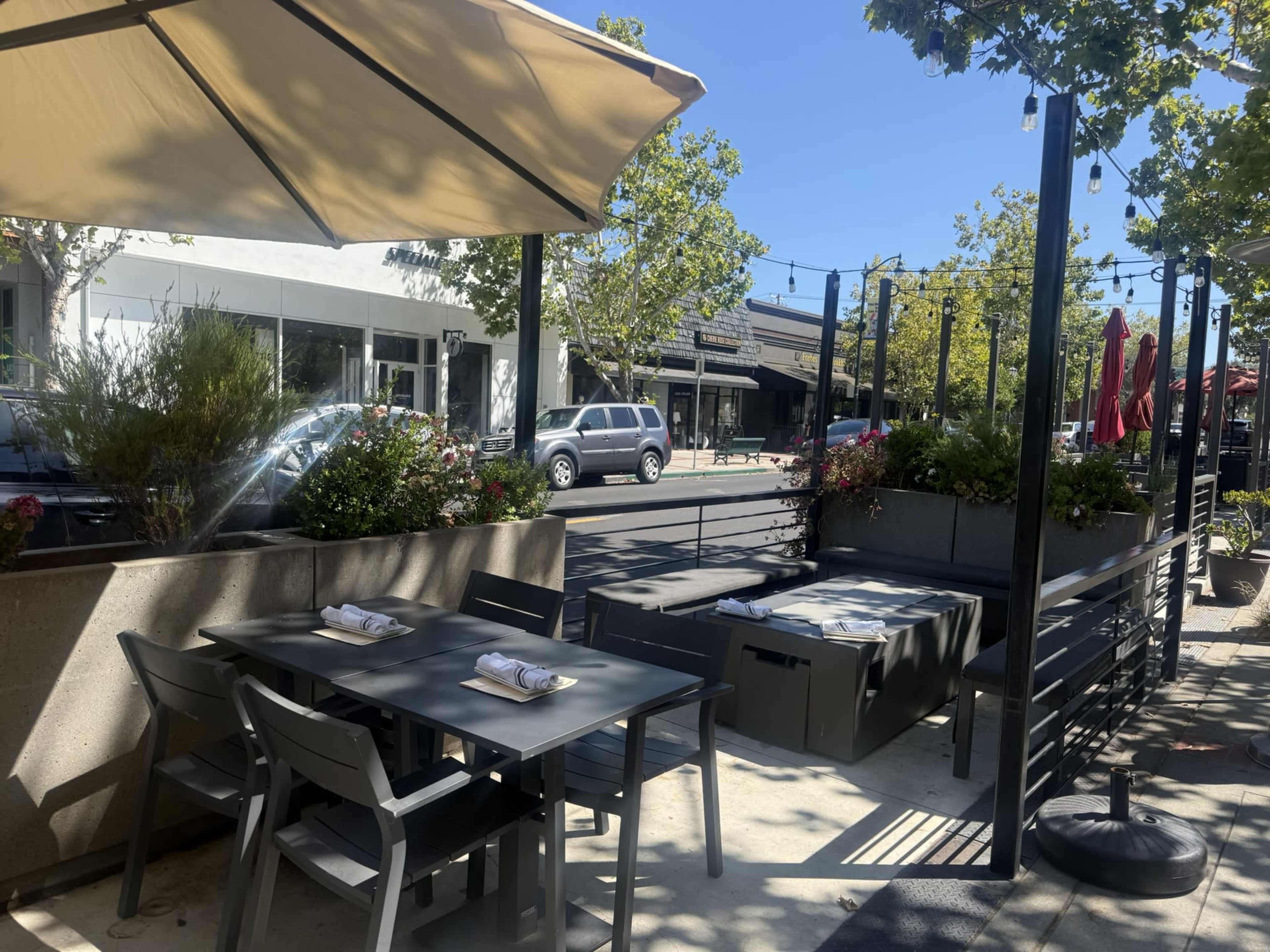The image shows a sidewalk dining area with tables and chairs shaded by an umbrella, flanked by planters and located near parked cars and storefronts.
