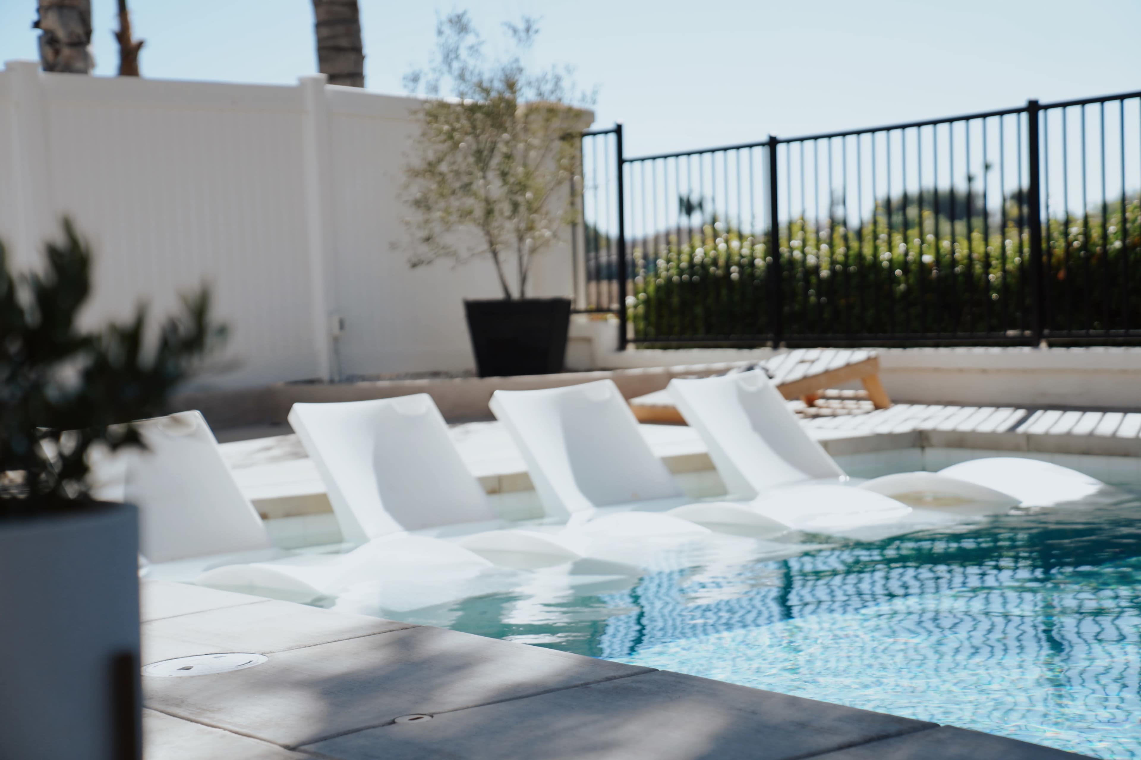 Lounge chairs are positioned partially submerged along the edge of a swimming pool, with a fence and greenery in the background.