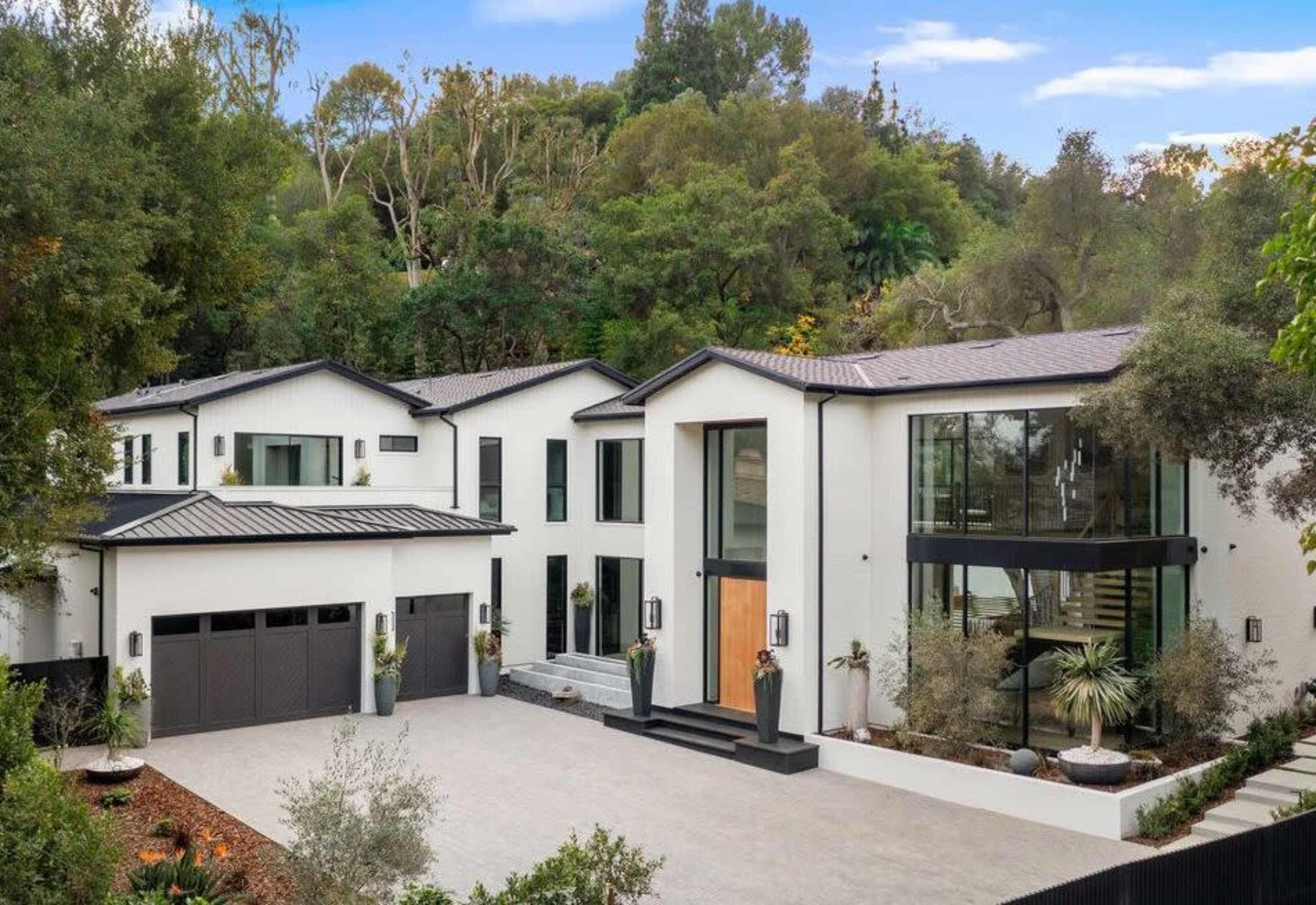 A modern two-story house with large windows, a symmetrical design, and a landscaped driveway is nestled among lush greenery.